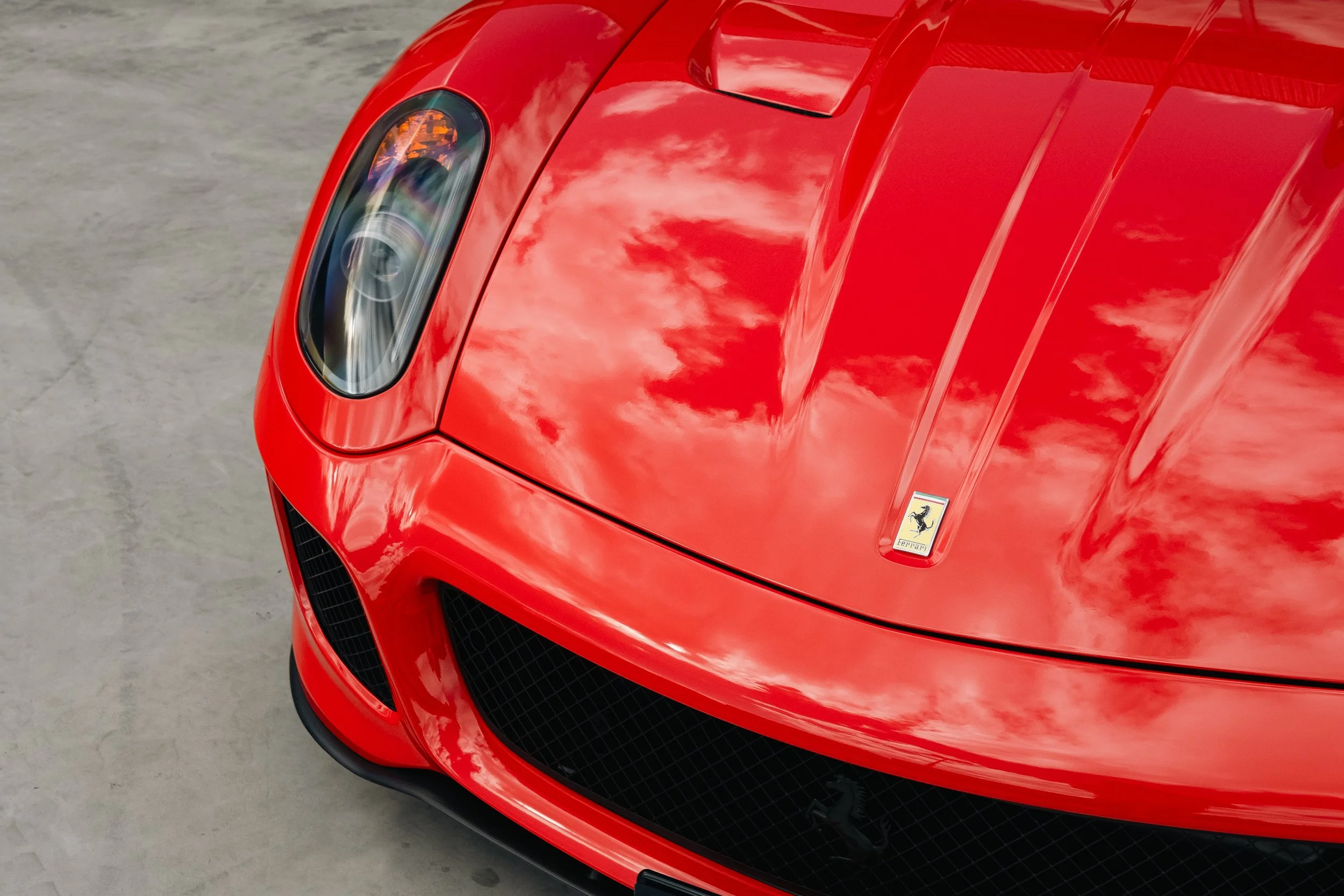 Close-up of the front of a red Ferrari sports car, showing the headlight, hood with Ferrari emblem, and front grille with the prancing horse logo.