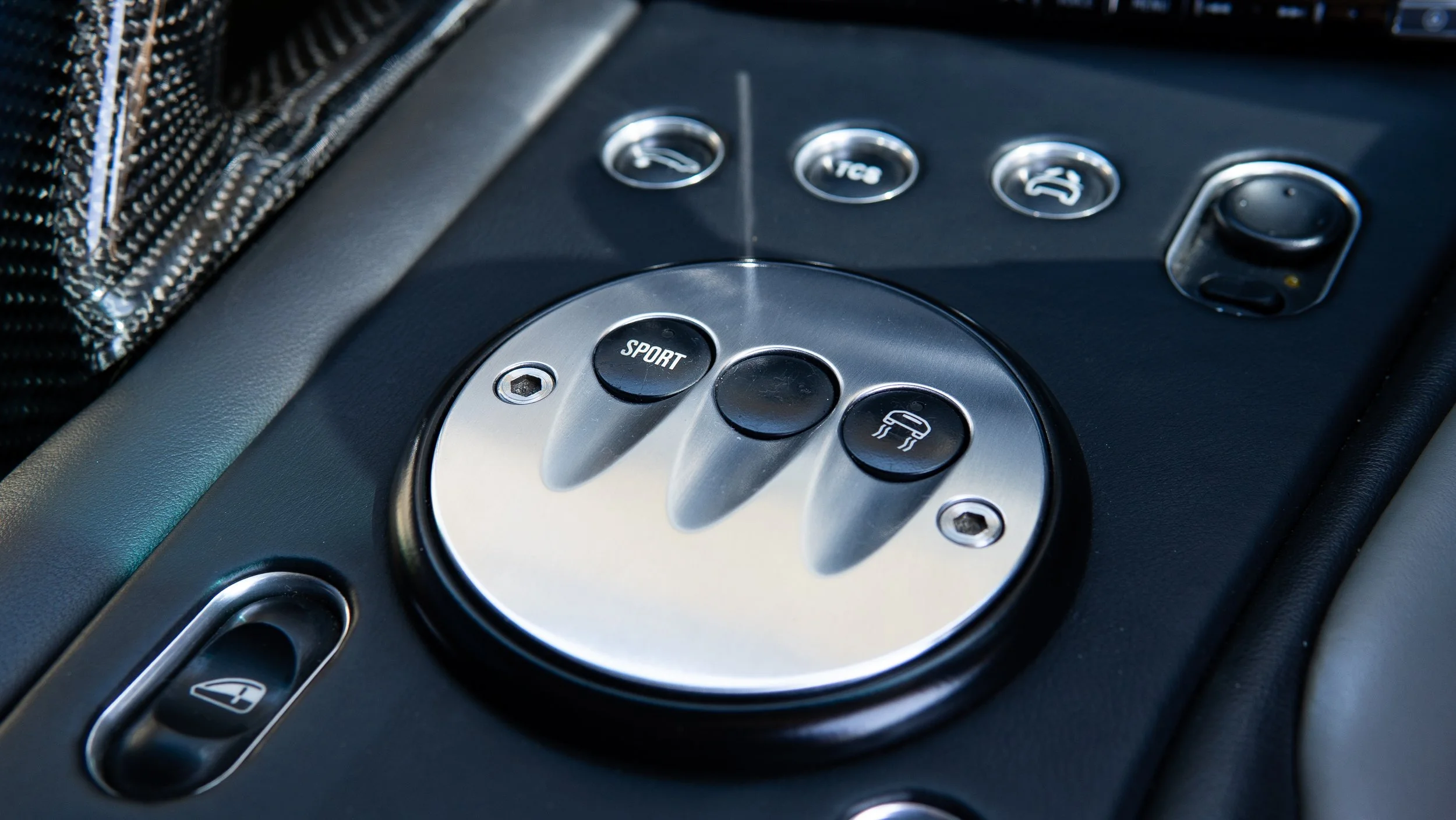 Close-up of a race car transmission control panel with metallic and black buttons labeled 'SPORT' and other icons, surrounding a circular area with various switches and indicators.