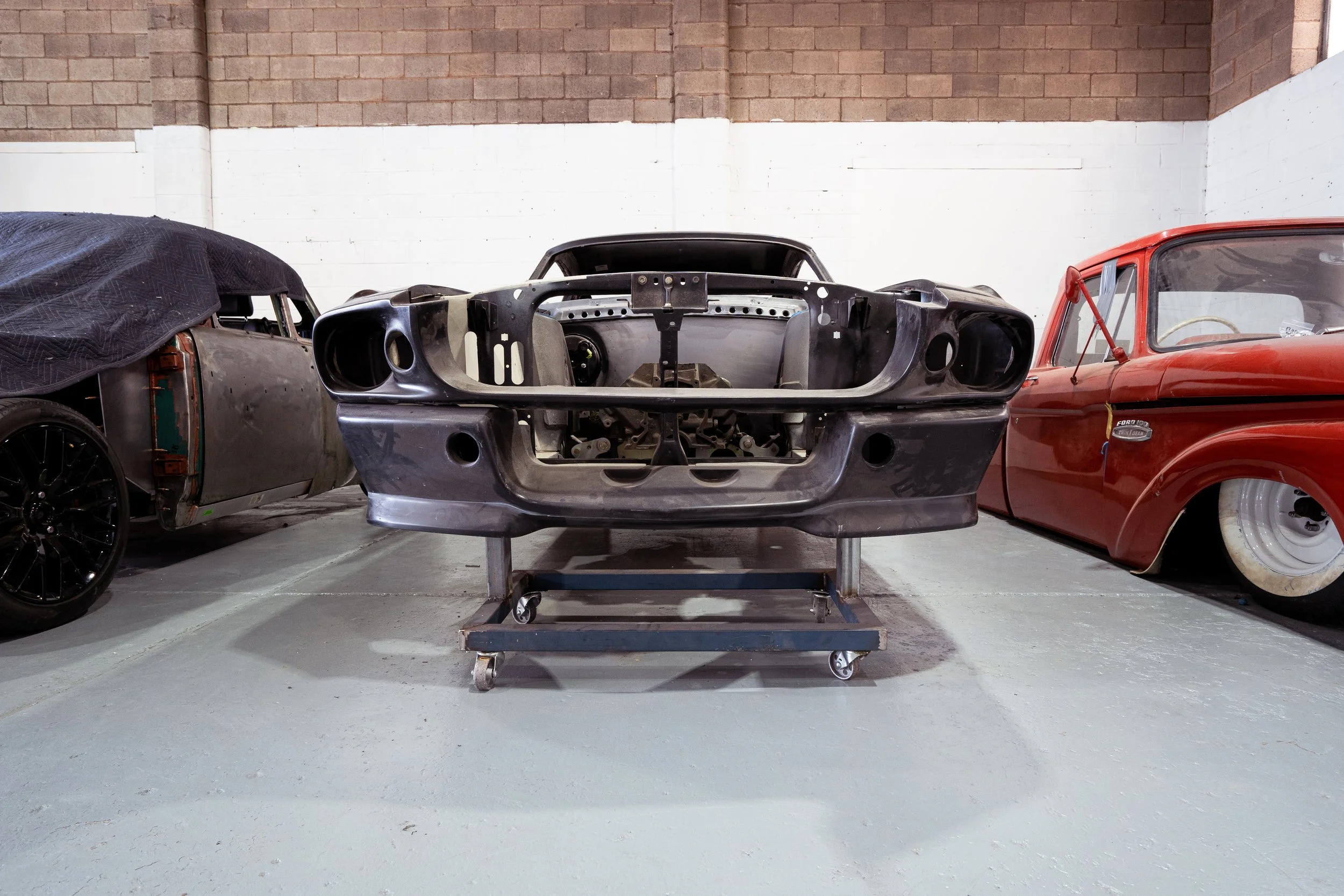 Car shell with no engine or interior parts on a wheeled cart, flanked by two other vintage cars in a garage or workshop.