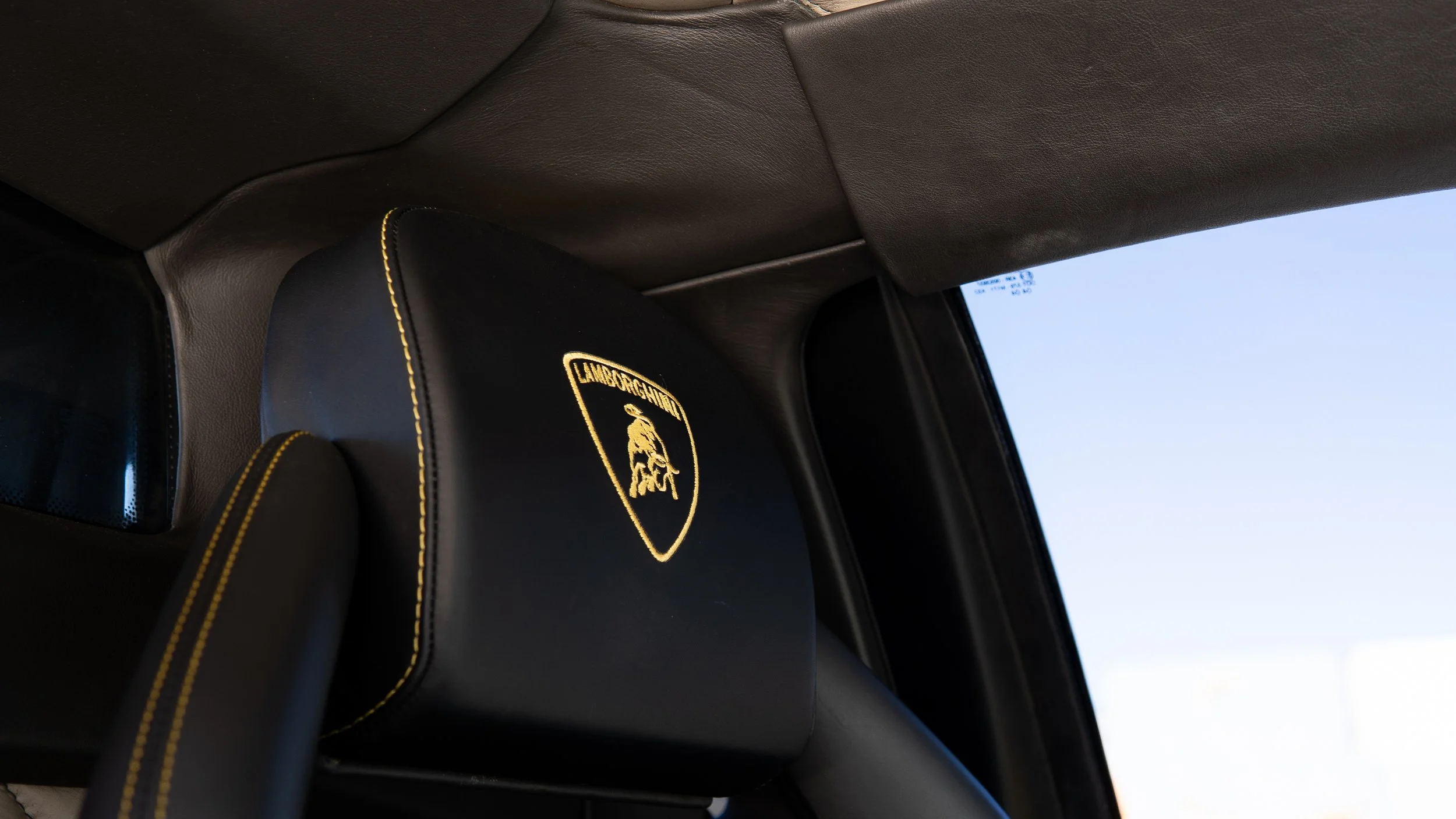 Interior view of a Lamborghini vehicle, showing a black headrest with the Lamborghini logo embroidered in gold thread, beige dashboard, and part of a window with a clear sky outside.