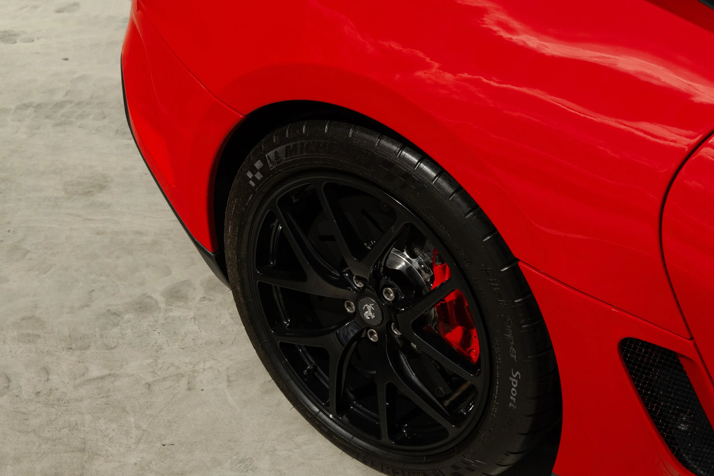 Close-up of a red sports car with a black wheel, tire, and brake caliper on a concrete surface.