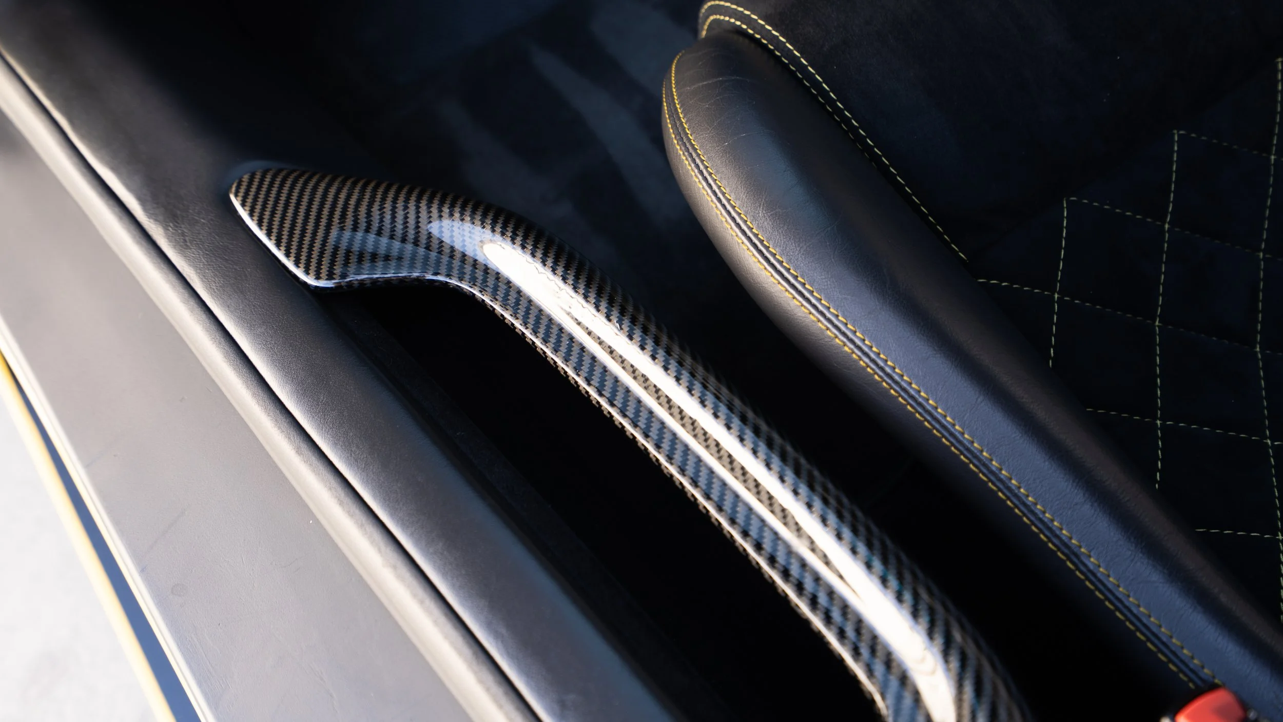 Close-up of a car interior showing a carbon fiber trim on the door and a black seat with yellow stitching.