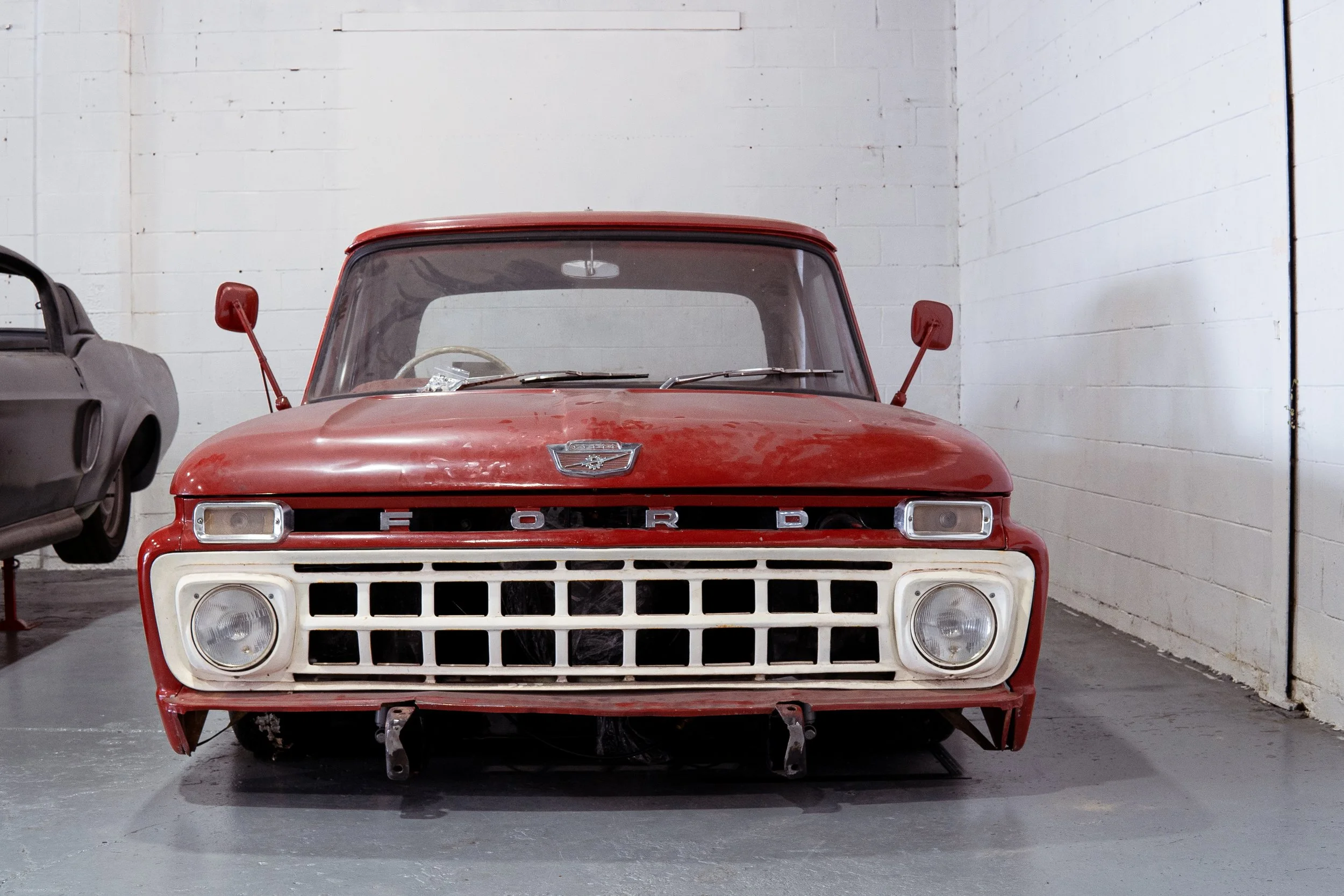 Red vintage Ford pickup truck inside a garage with white brick walls, with a black car partially visible to the left.