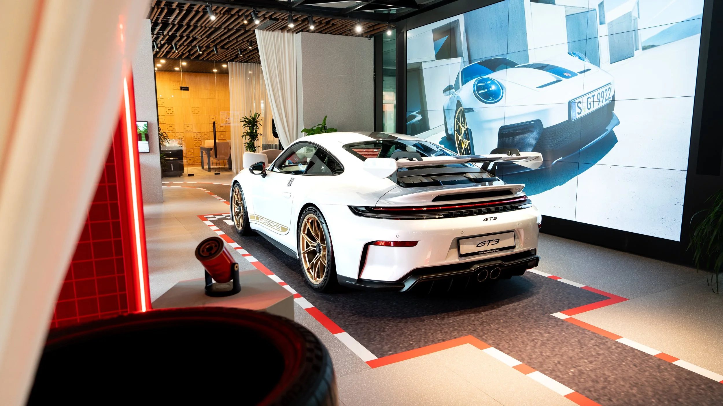 A white Porsche 911 GT3 sports car with gold rims on display inside a showroom, with a large screen behind it showing a similar car from the front.