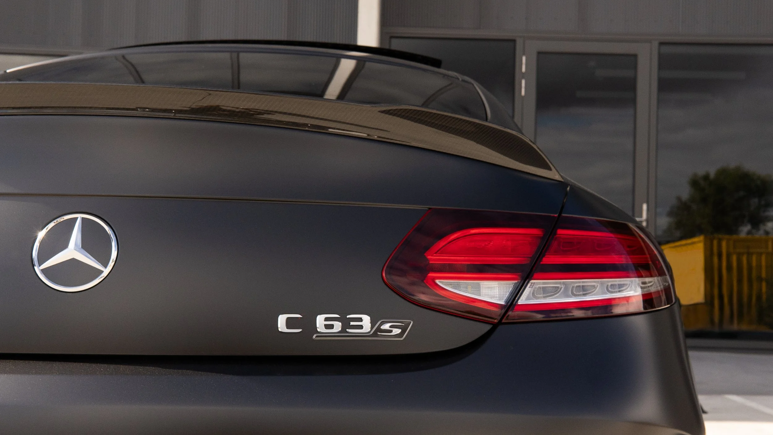 Close-up of the rear of a black Mercedes-Benz C 63 S with the Mercedes logo, taillights, and model badge, parked outside a building with glass doors and windows.