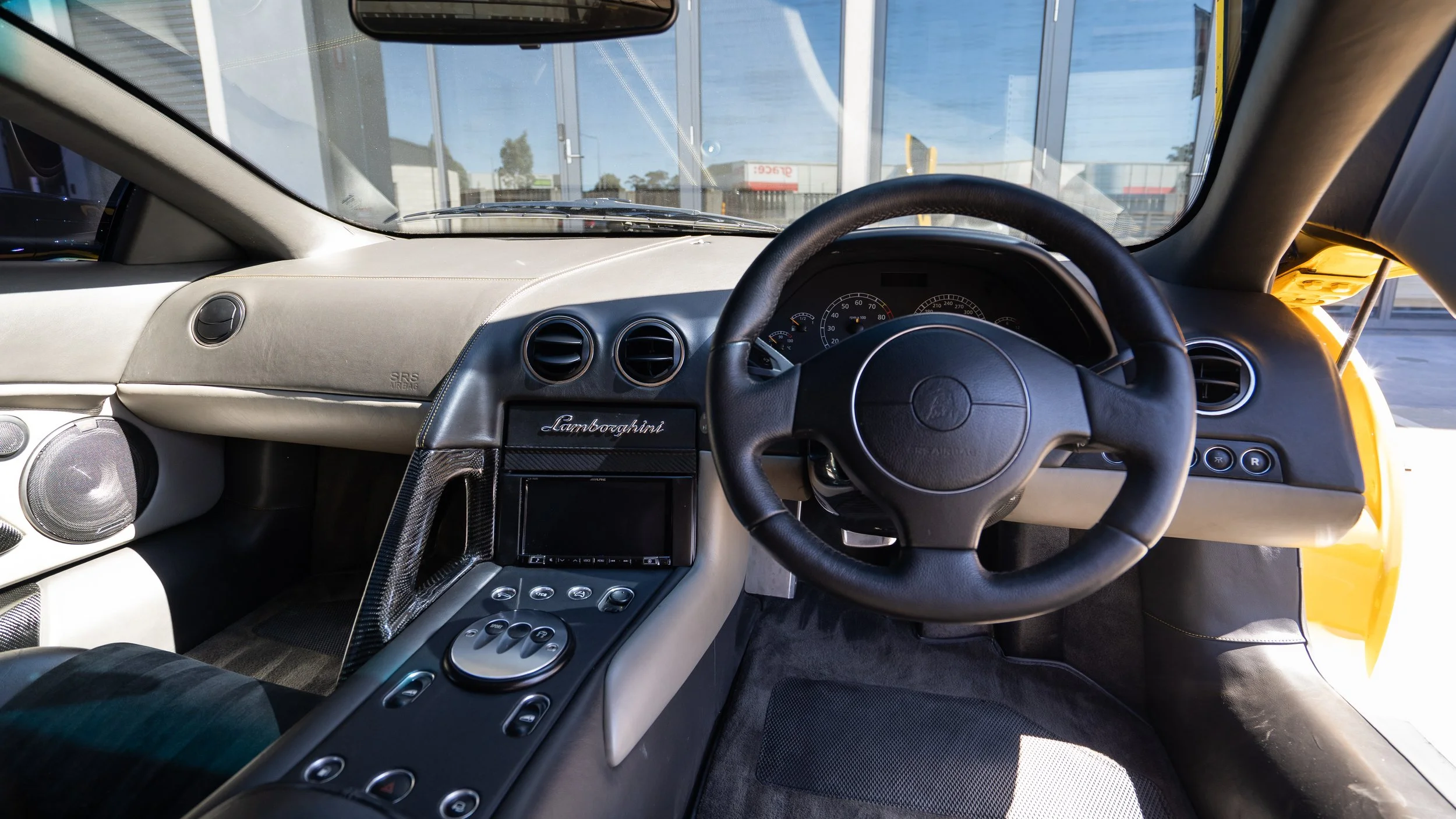 View of a Lamborghini car interior showing the dashboard, steering wheel, and center console with controls, with a yellow exterior visible through the windshield.