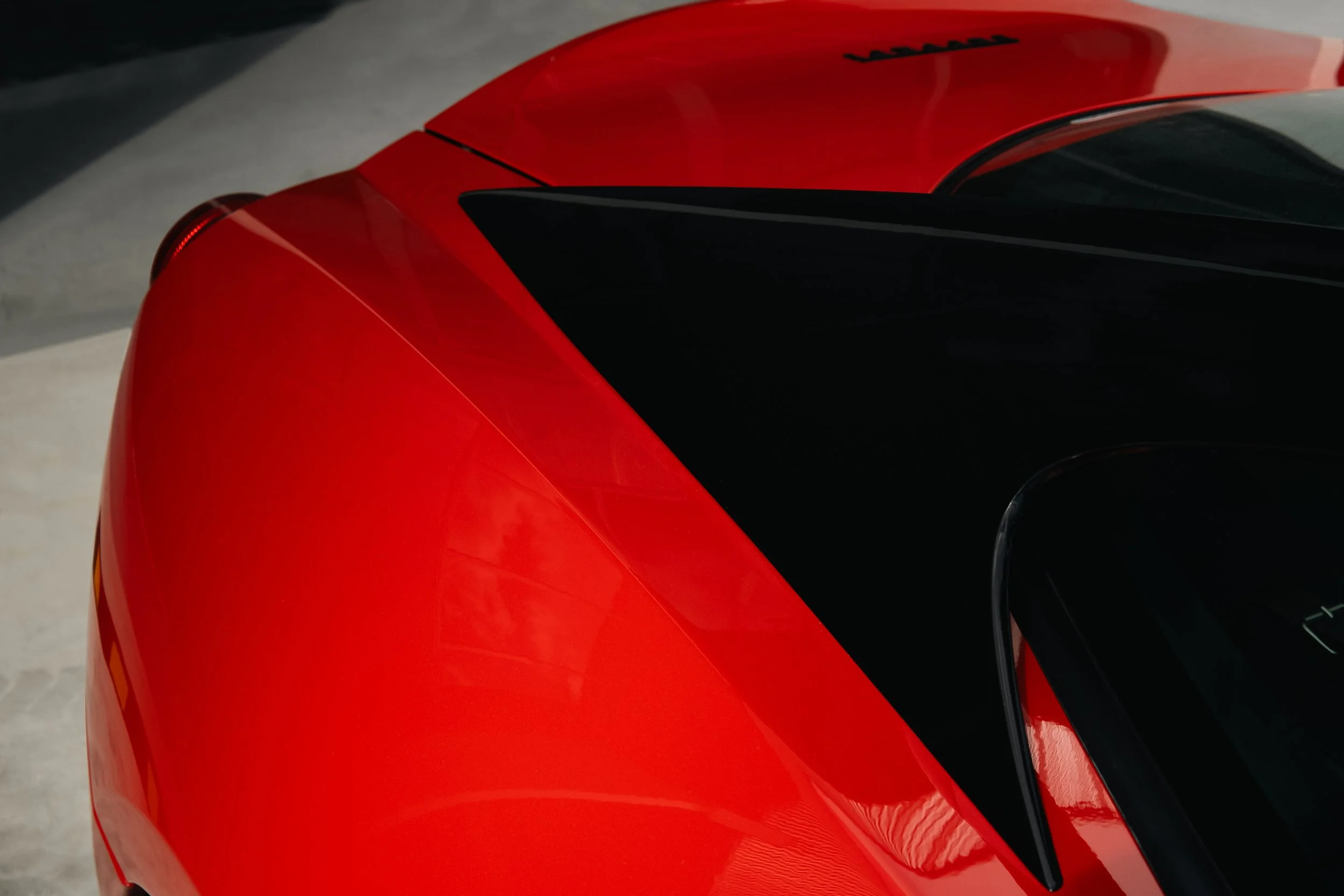 Close-up of the rear end of a red sports car with a black roof and spoiler, parked on a light-colored surface.