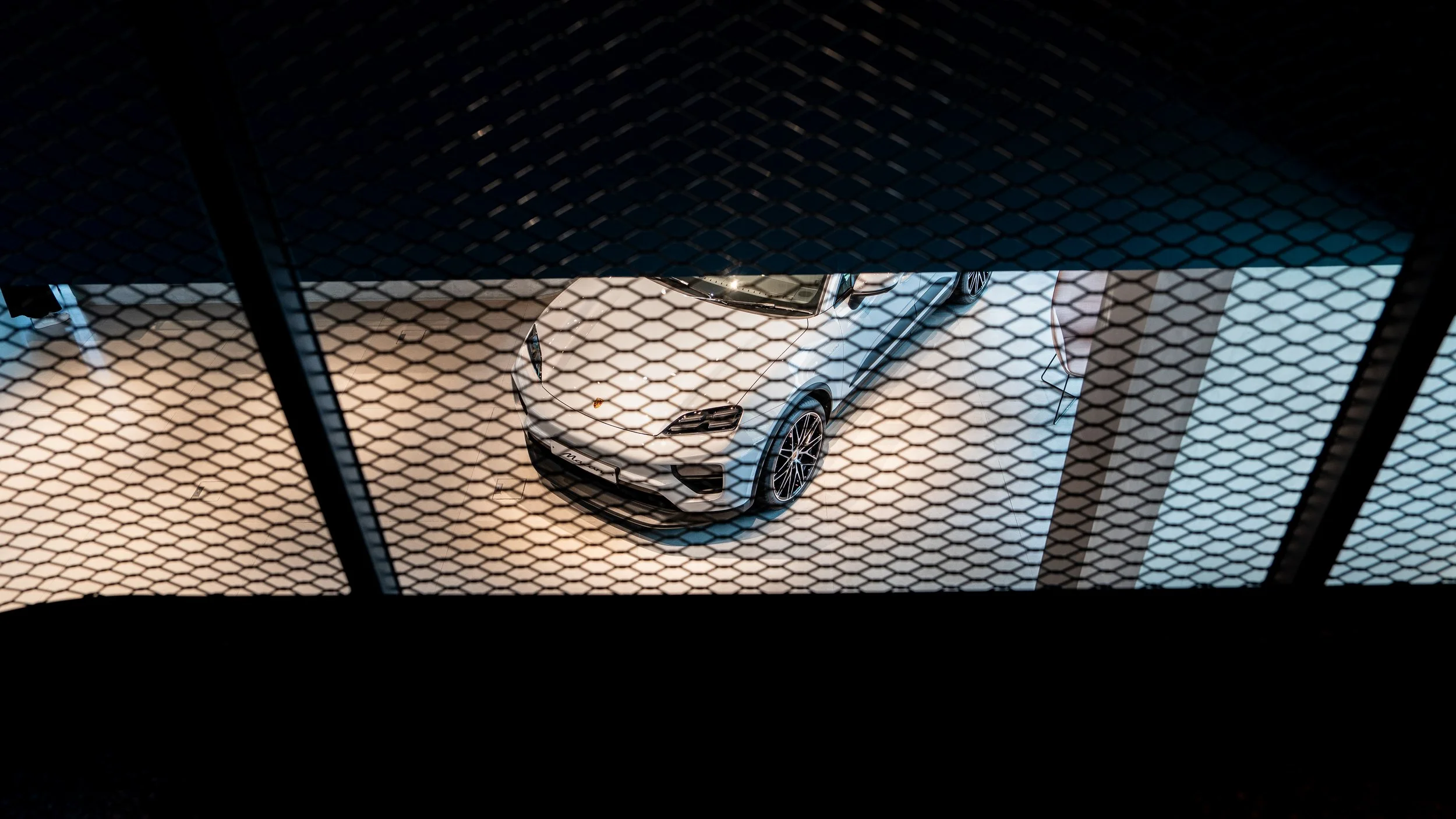 View of a white car through a metal mesh barrier from above.