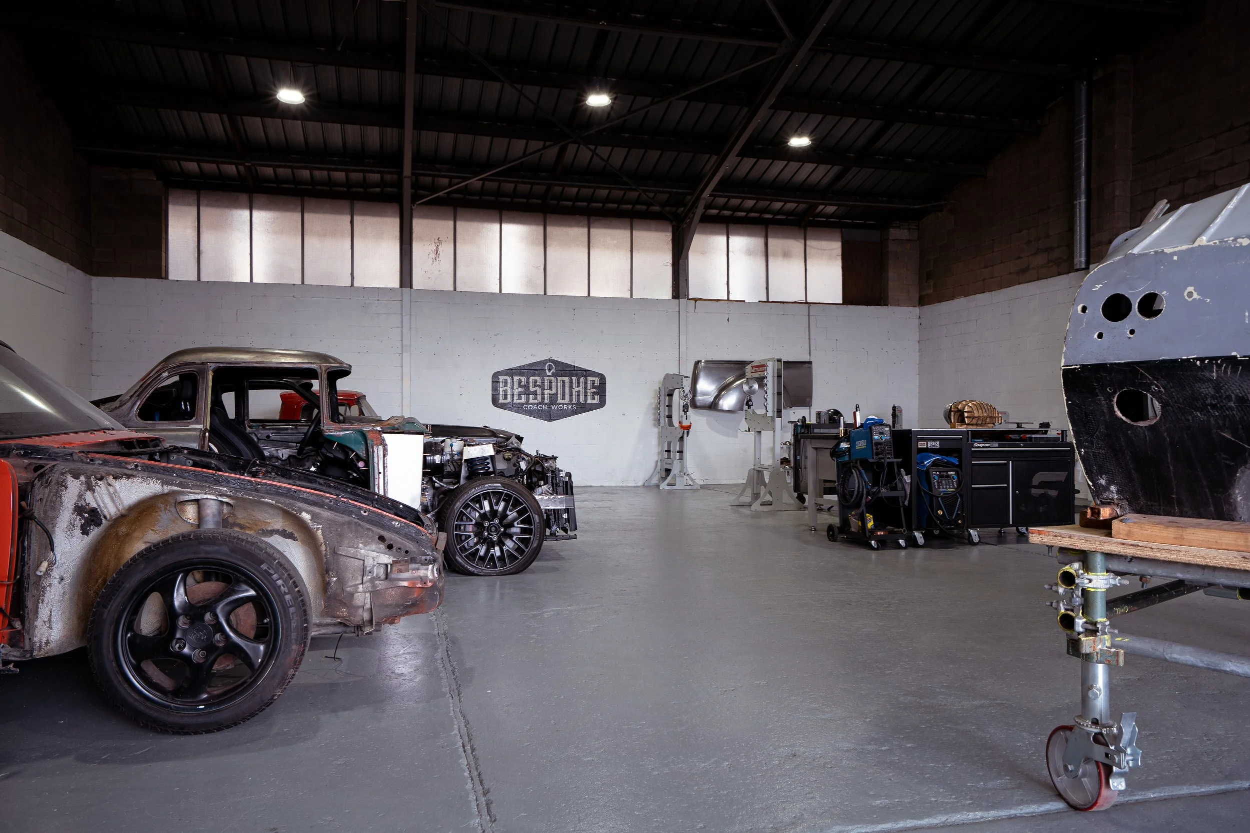 An auto repair workshop with partially disassembled cars, tools, and equipment, including a rusted car and a large metal work table.