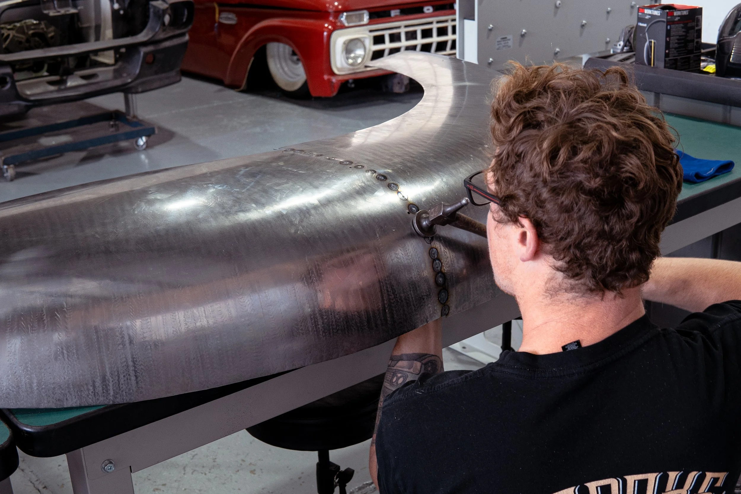 A person with curly hair and glasses working on a large metal surface with a welding or grinding tool, in a workshop with car parts in the background.