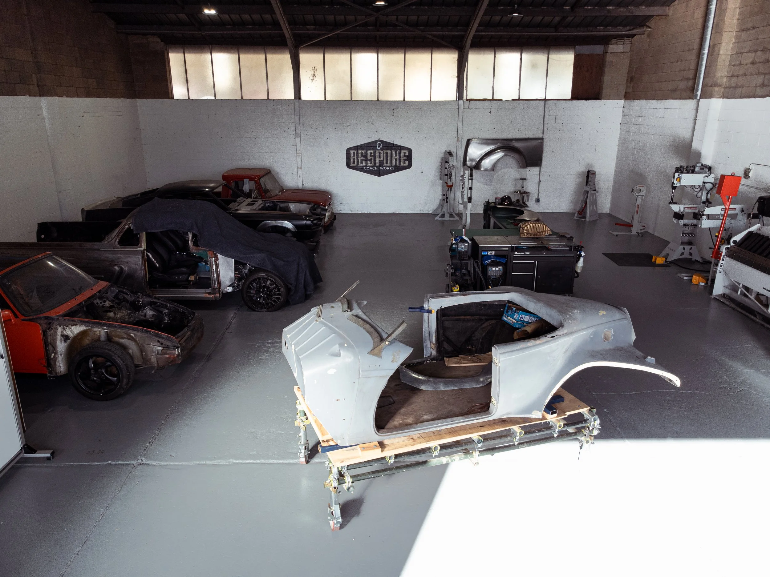 An indoor garage or workshop with car parts, tools, and partially assembled cars. Several vehicles in various stages of restoration or repair are visible, including a clear car body on a stand in the foreground and other cars covered with protective sheets.