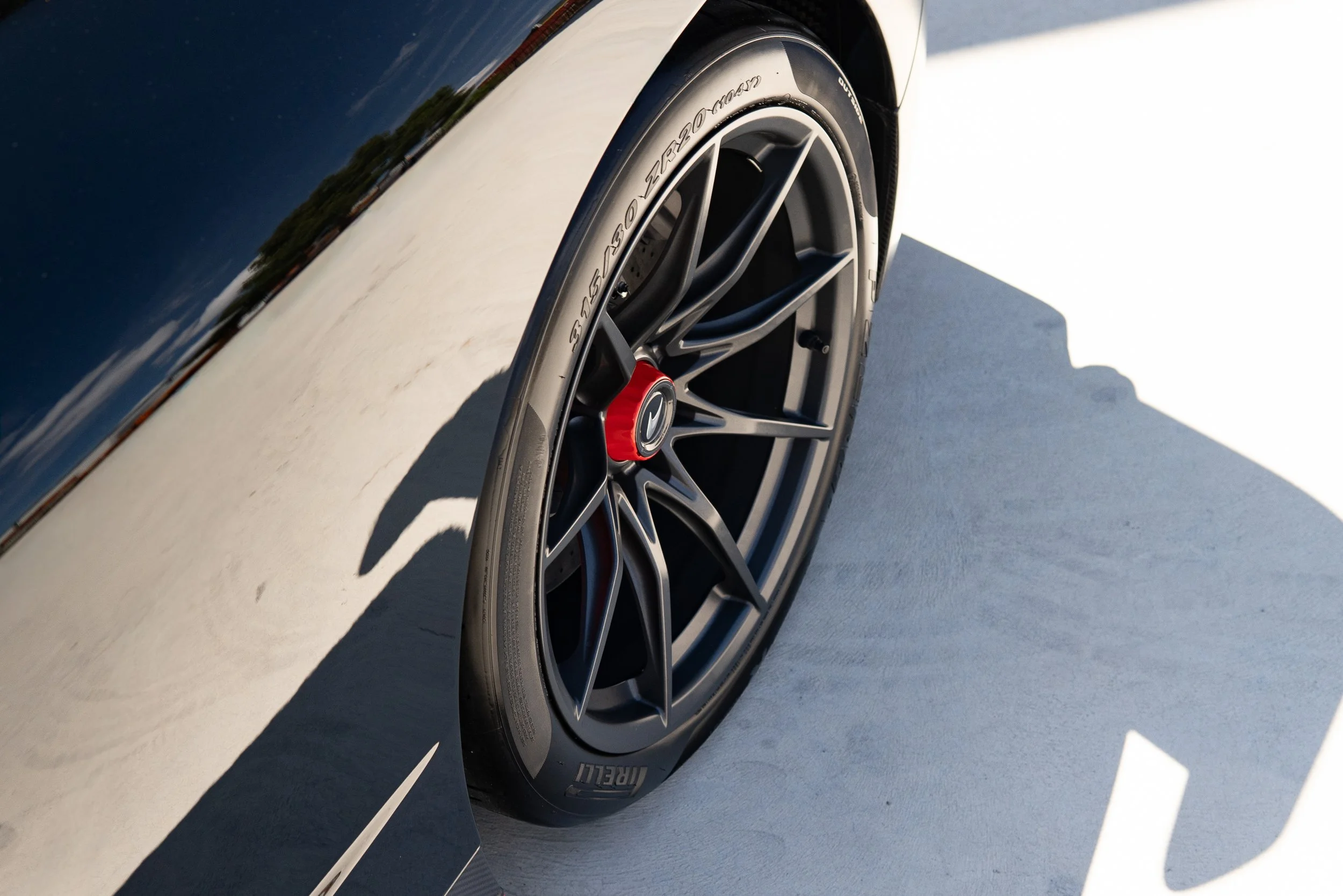 A close-up of a car wheel with a black alloy rim and a Pirelli tire on a light-colored surface, with a shadow cast to the right.
