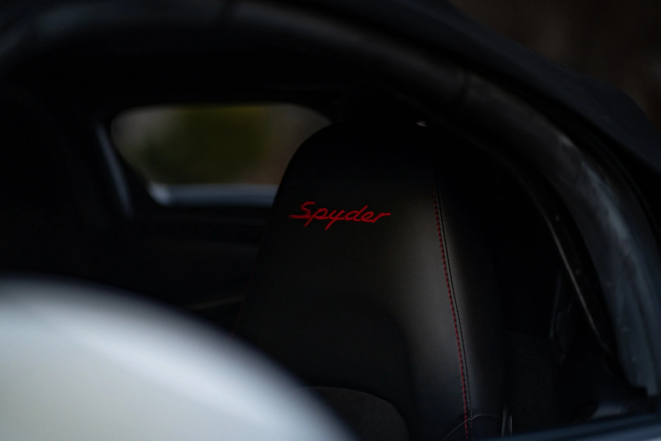 The photo shows the interior of a vehicle, focusing on a black leather car seat with red stitching and the word 'Spyder' embroidered in red on the headrest. The background appears blurry with the vehicle's window visible.