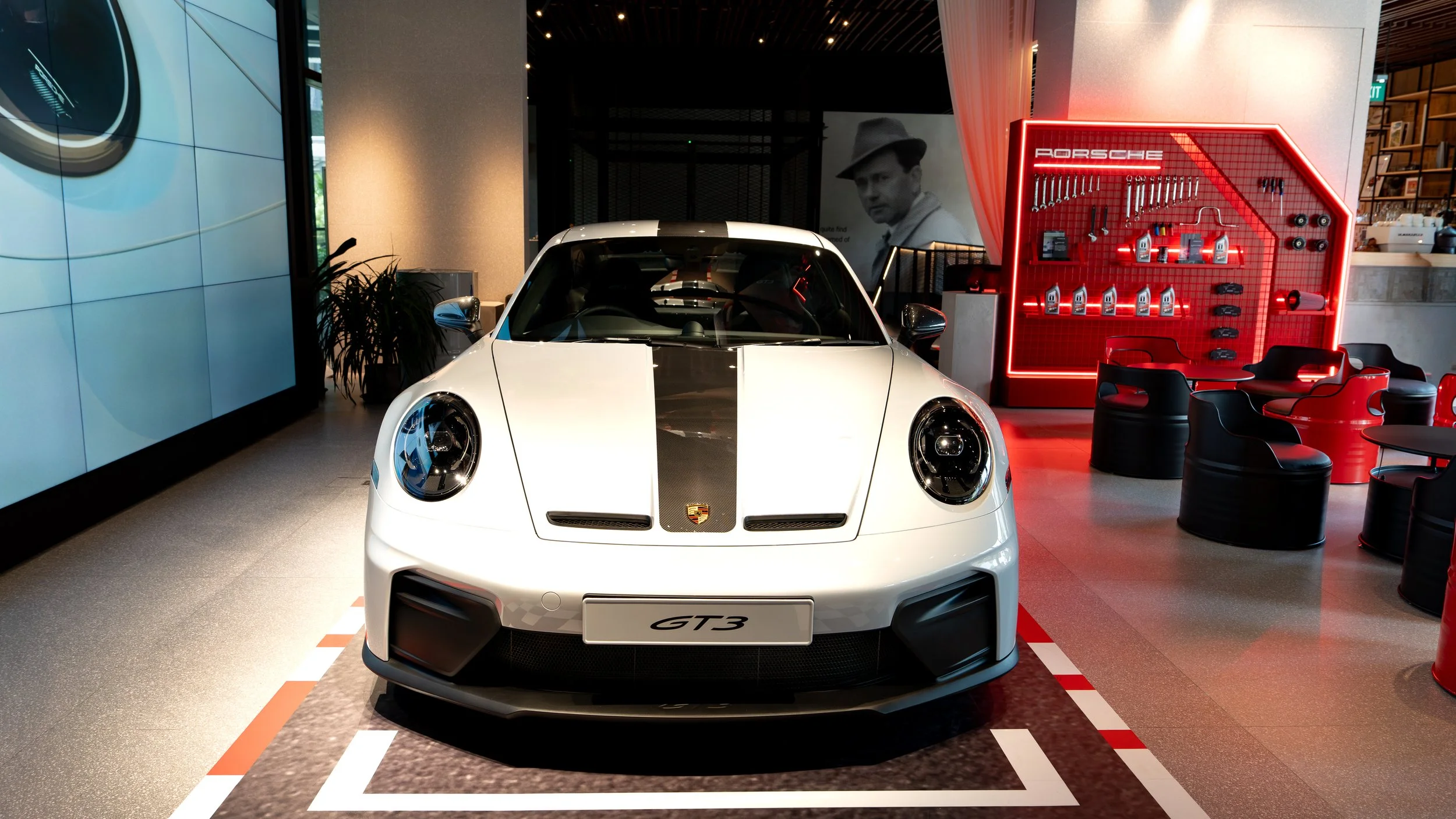 White Porsche 911 GT3 sports car displayed indoors, with a black stripe on the hood, in front of a red Porsche-themed tool display and seating area.