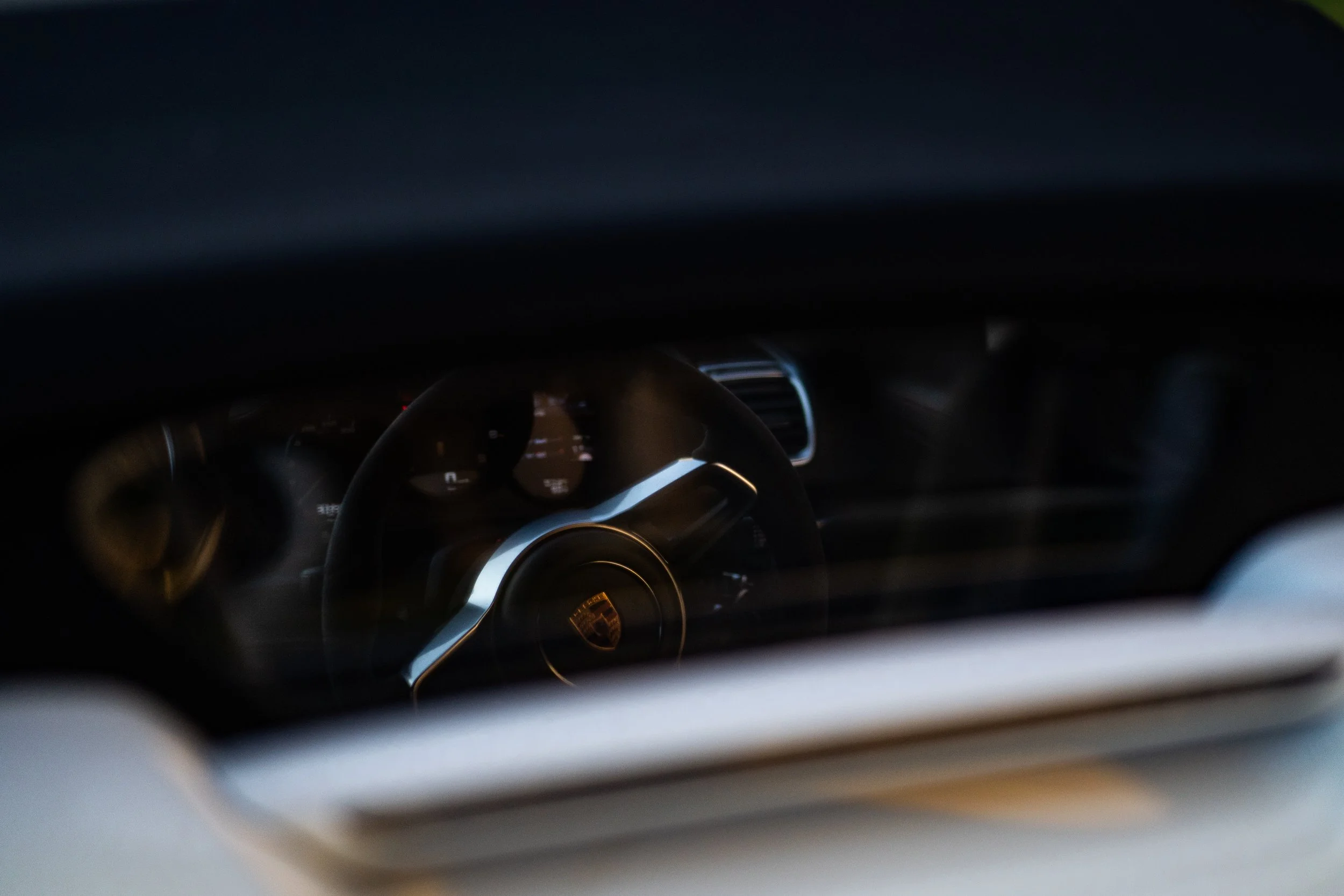Close-up of a car's interior dashboard viewed through the windshield, featuring a Porsche emblem on the steering wheel with gauges faintly visible in the background.