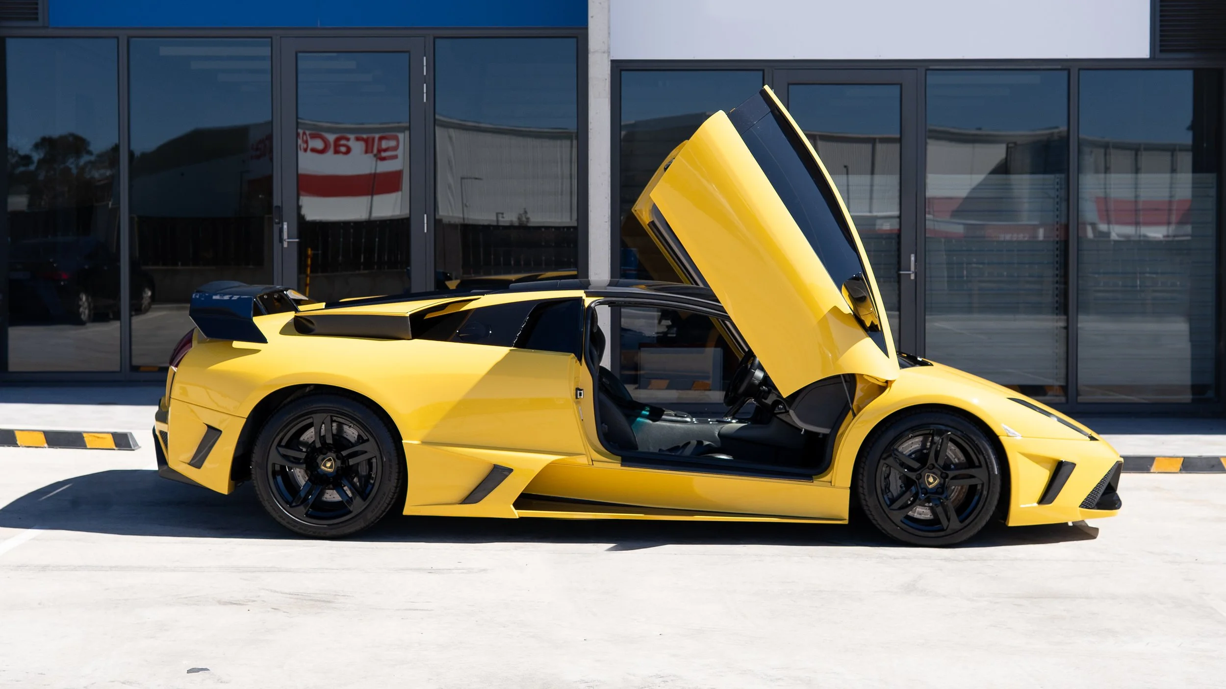 A yellow sports car with its door and hood open, parked outside a modern building with glass windows.