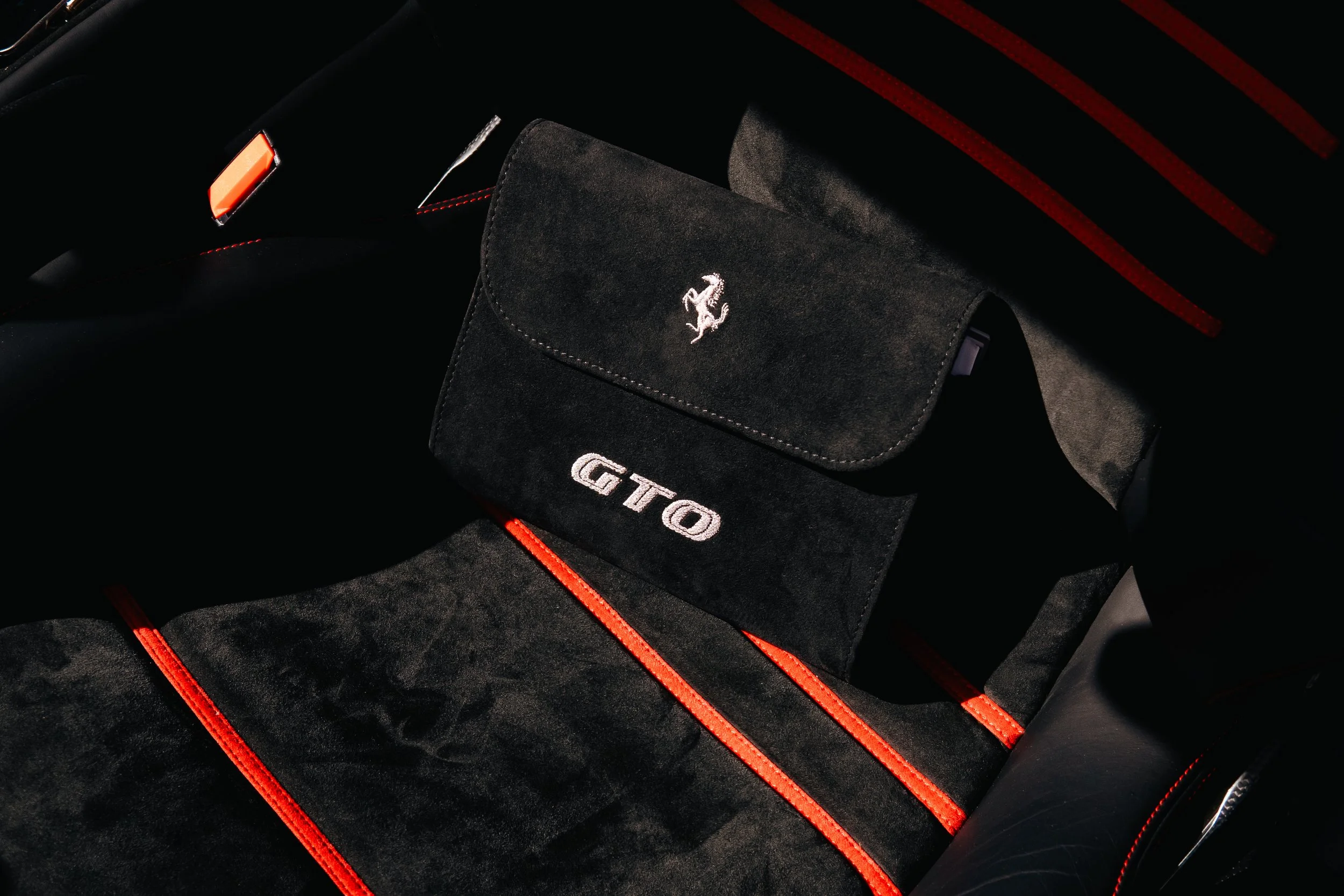 Black car seat with red stitching, a black suede pouch with a white Ferrari logo and the letters 'GTO' embroidered on it.