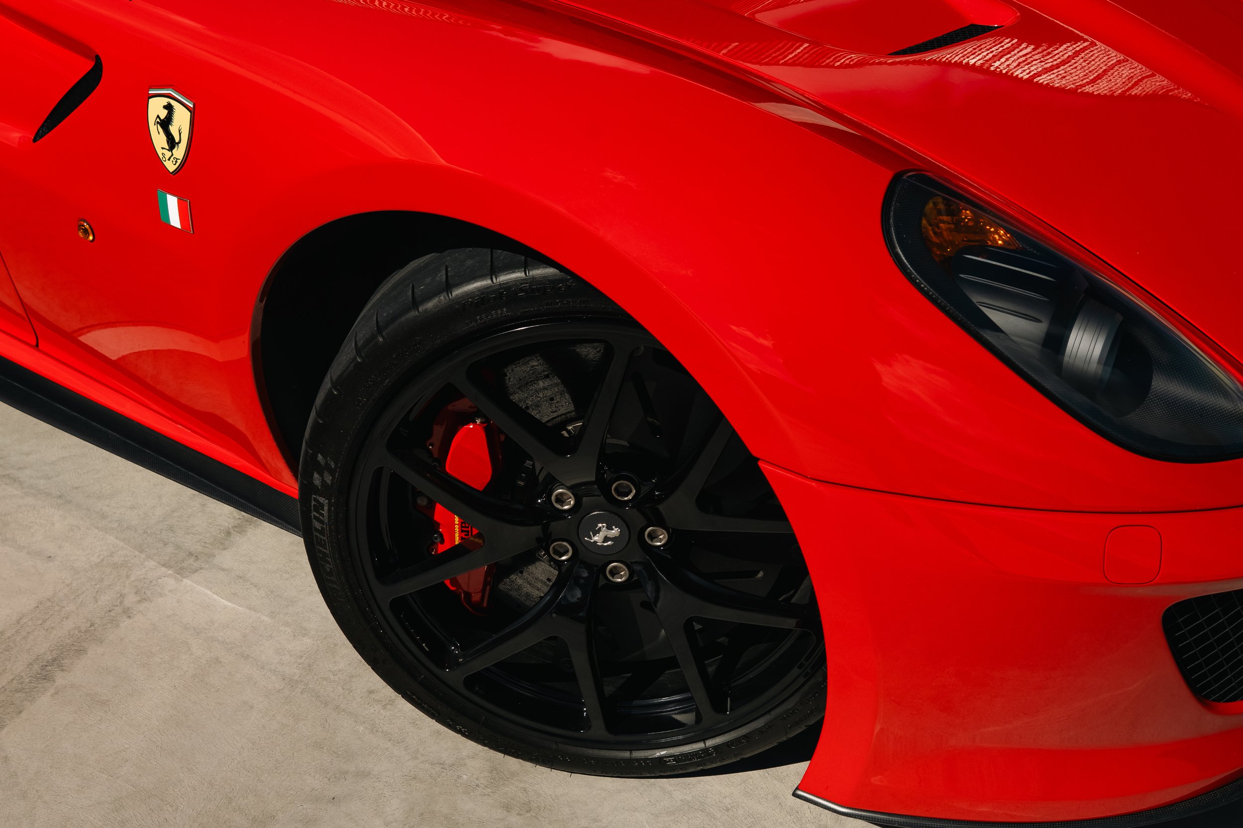 Close-up of a red Ferrari sports car, showing the front wheel, black rims, and brake caliper, with the Ferrari logo and Italian flag emblem on the side.