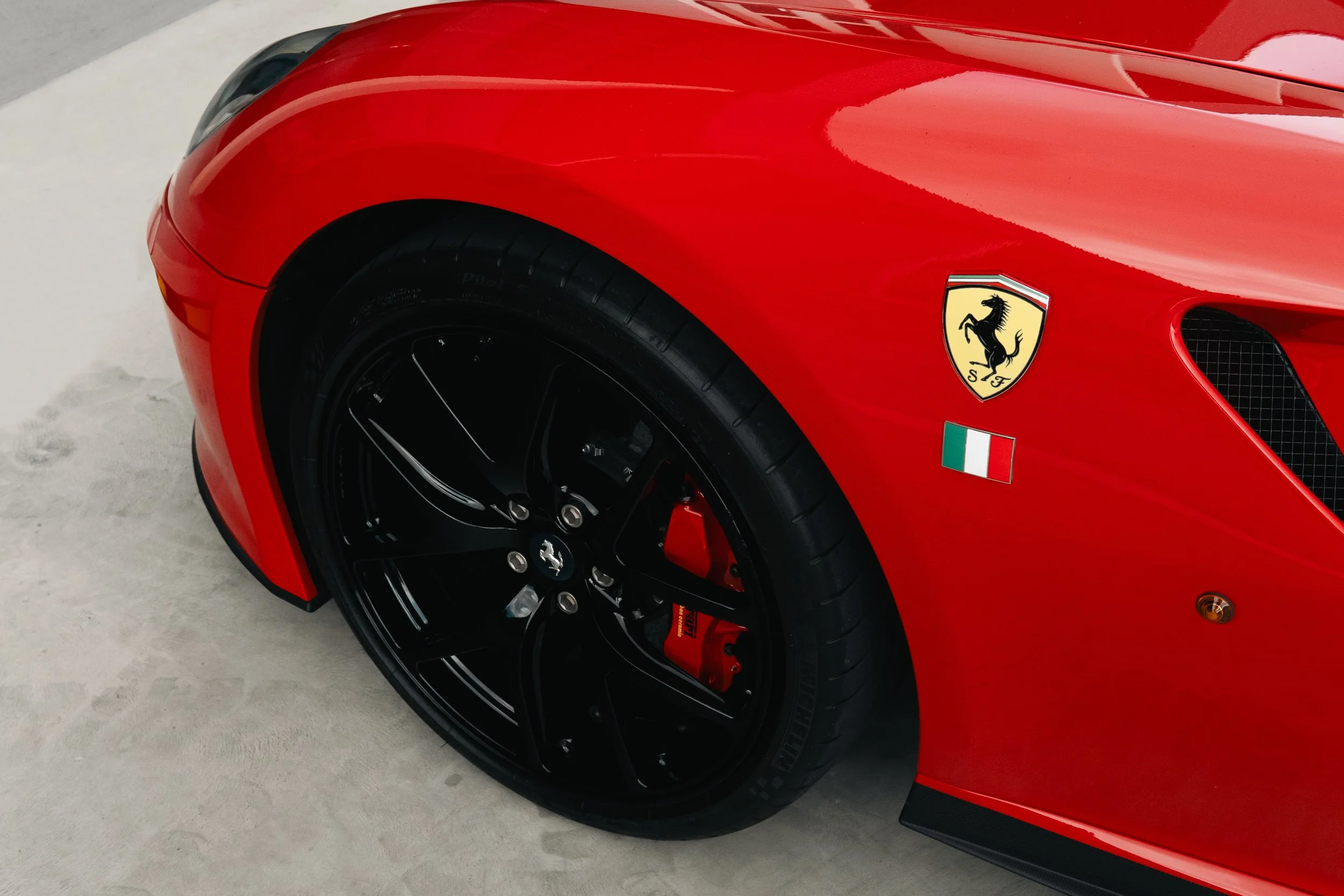 Close-up of the front section of a red Ferrari sports car featuring a black wheel, Ferrari emblem, and an Italian flag sticker.