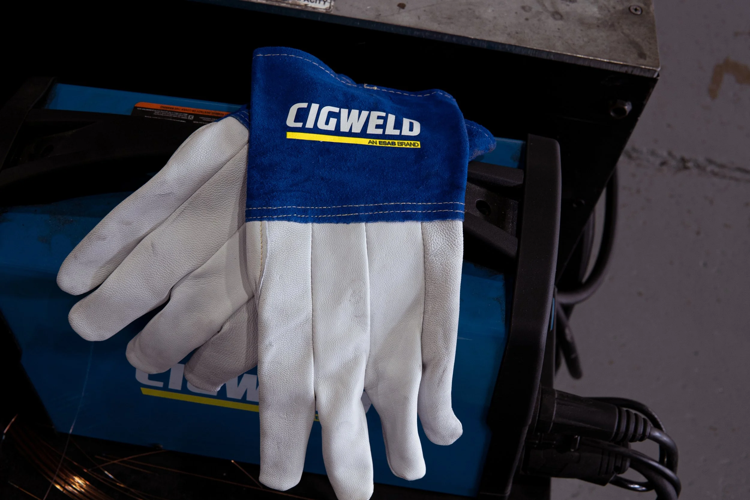 A pair of white work gloves with a blue wristband, labeled 'CIGWELD' and 'AN ESAB BRAND', resting on a blue cardboard box on a black cart, with a gray concrete floor in the background.