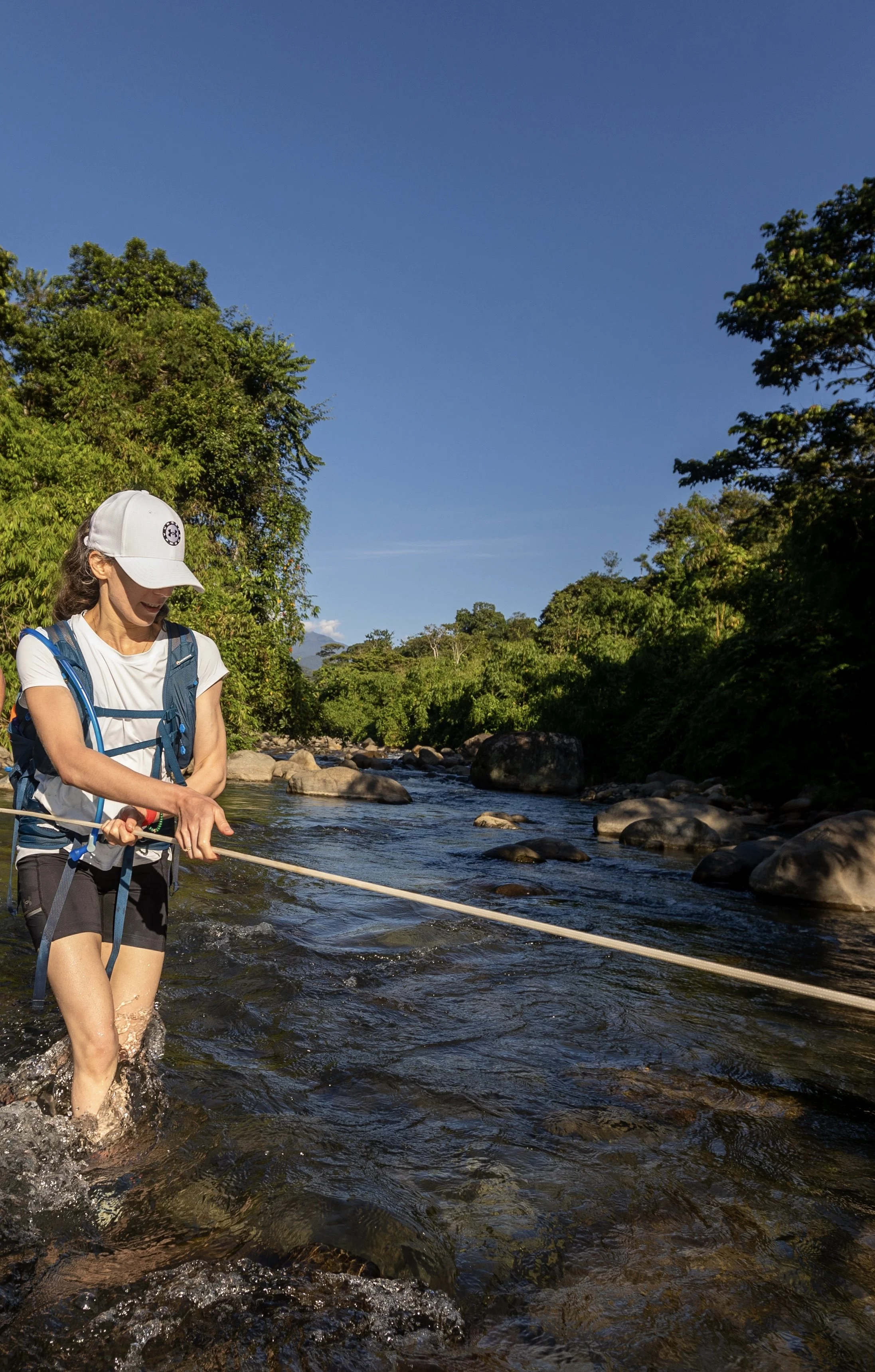 A woman wearing a white cap, white t-shirt, backpack, and black shorts stands in a river, holding a rope crossing a river with a lush green forest in the background under a clear blue sky.