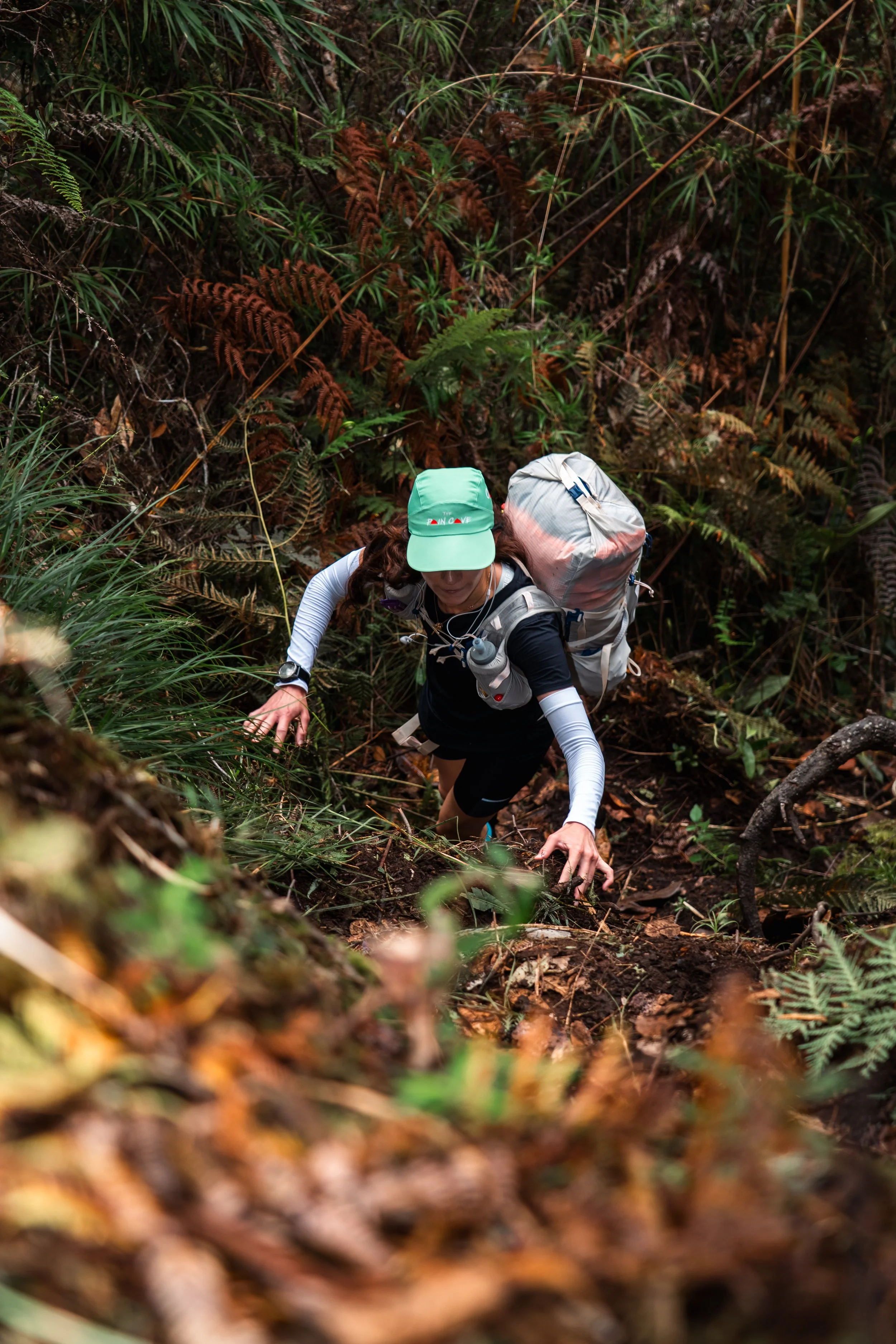 A person climbing a steep, muddy trail in a dense forest, equipped with a backpack, wearing a cap, and using their hands to assist in climbing.