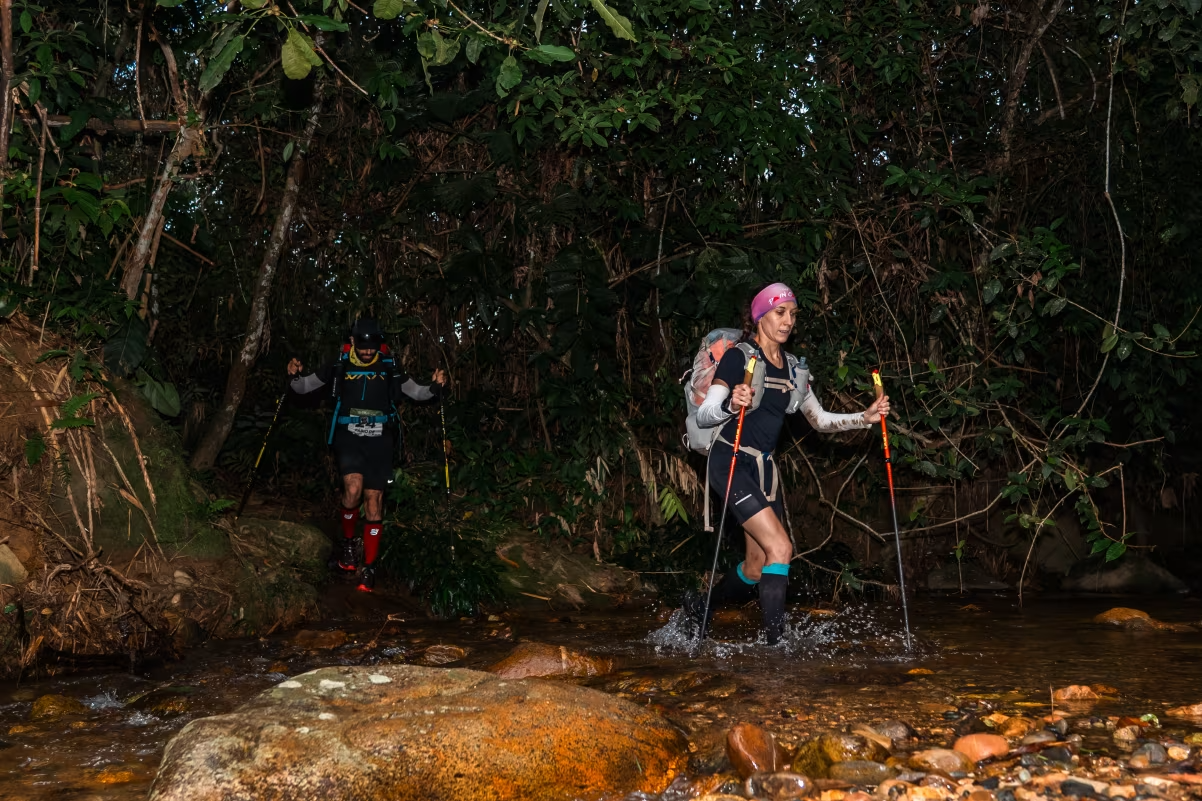 Two hikers crossing a shallow stream in a dense forest, using trekking poles and wearing backpacks.