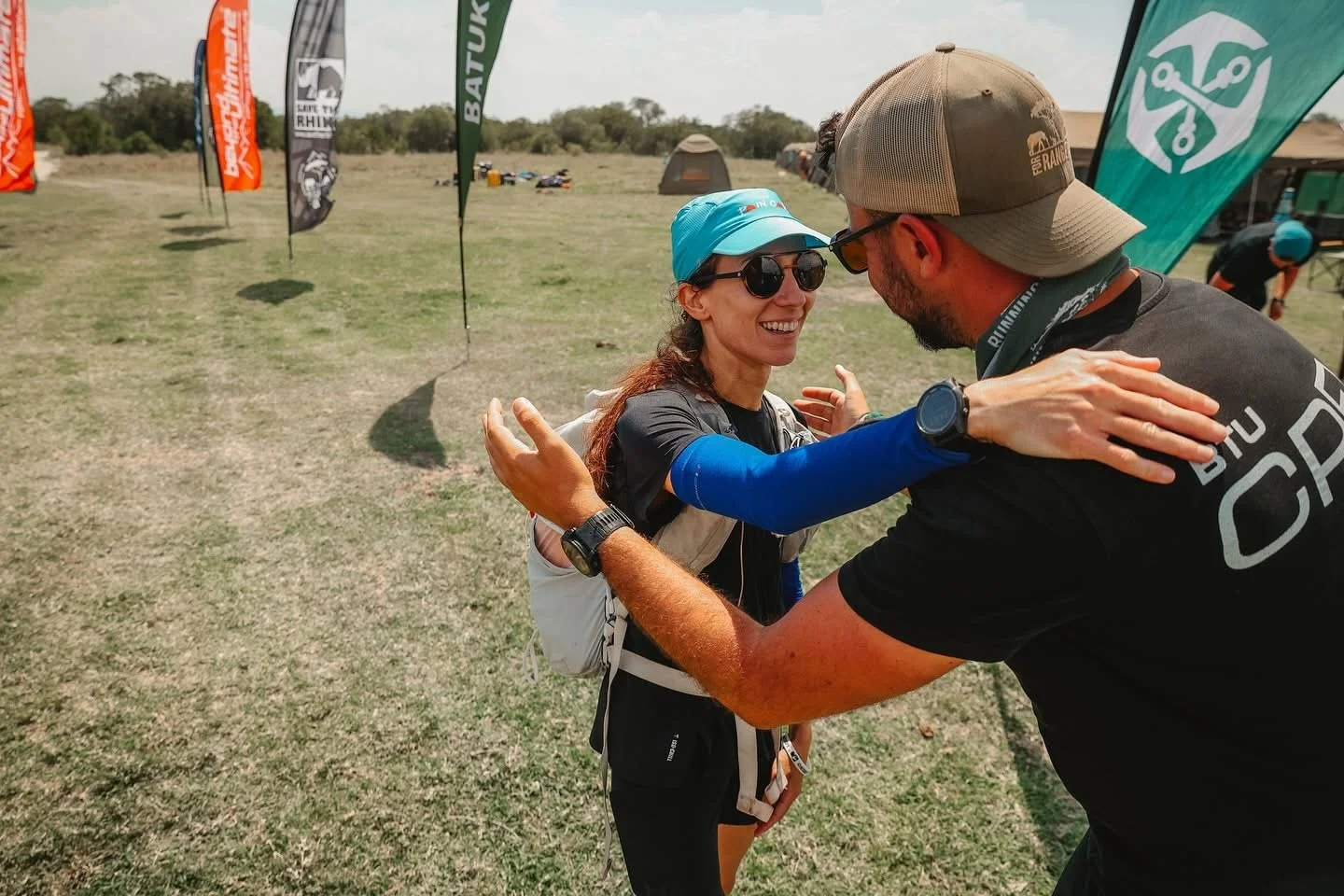 A woman with sunglasses and a blue cap hugging a man with a baseball cap and sunglasses at an outdoor event with flags and tents in the background.