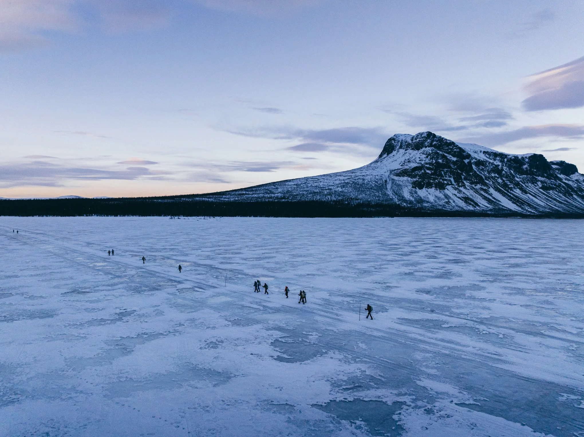 A group of people walking across a frozen lake with snow-covered mountains in the background during dusk.