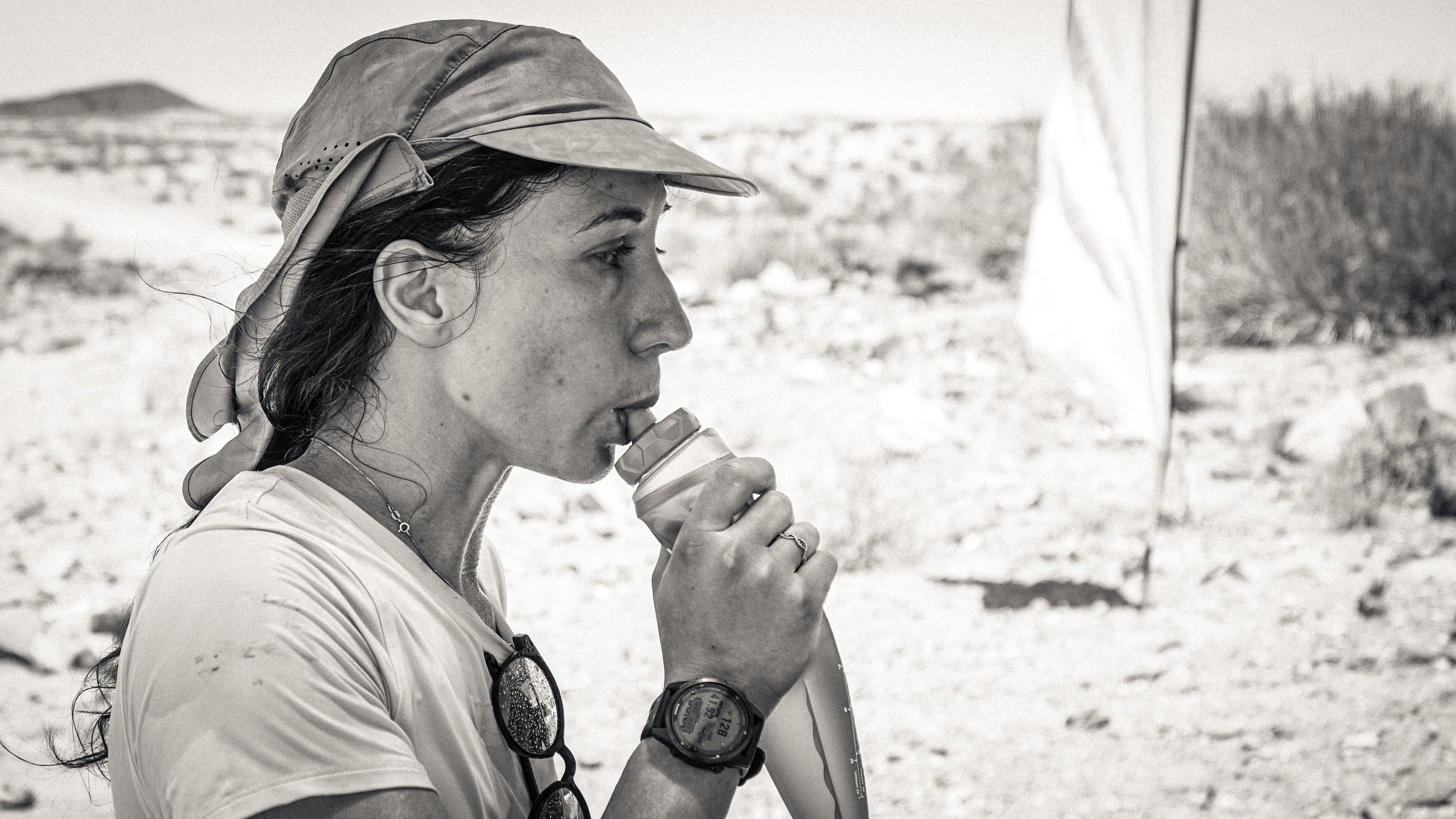 A woman in outdoors in a desert environment with a hat, drinking from a water bottle. Race. Athlete.