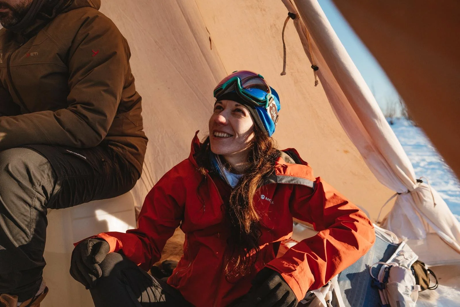 A woman smiling, wearing a red jacket, blue beanie, and spy goggles, sitting inside a tent during winter