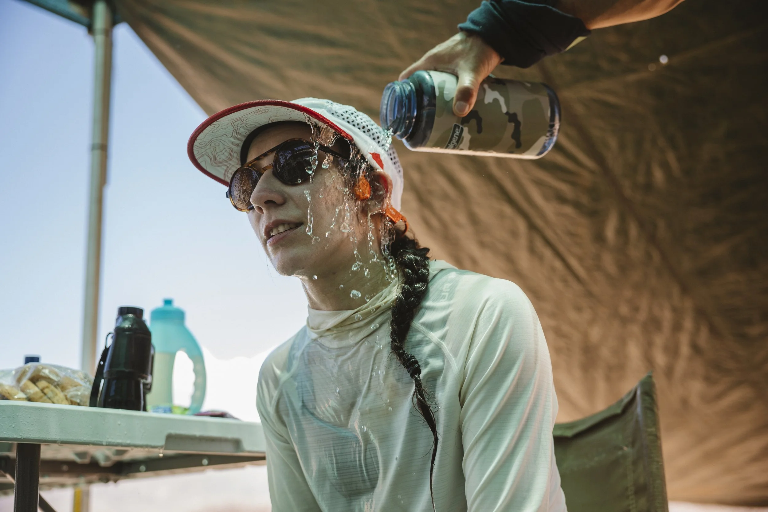 A woman wearing sunglasses and a wide-brimmed hat gets water poured on her head from a camouflage-patterned water bottle while sitting outdoors at a table under a canopy.