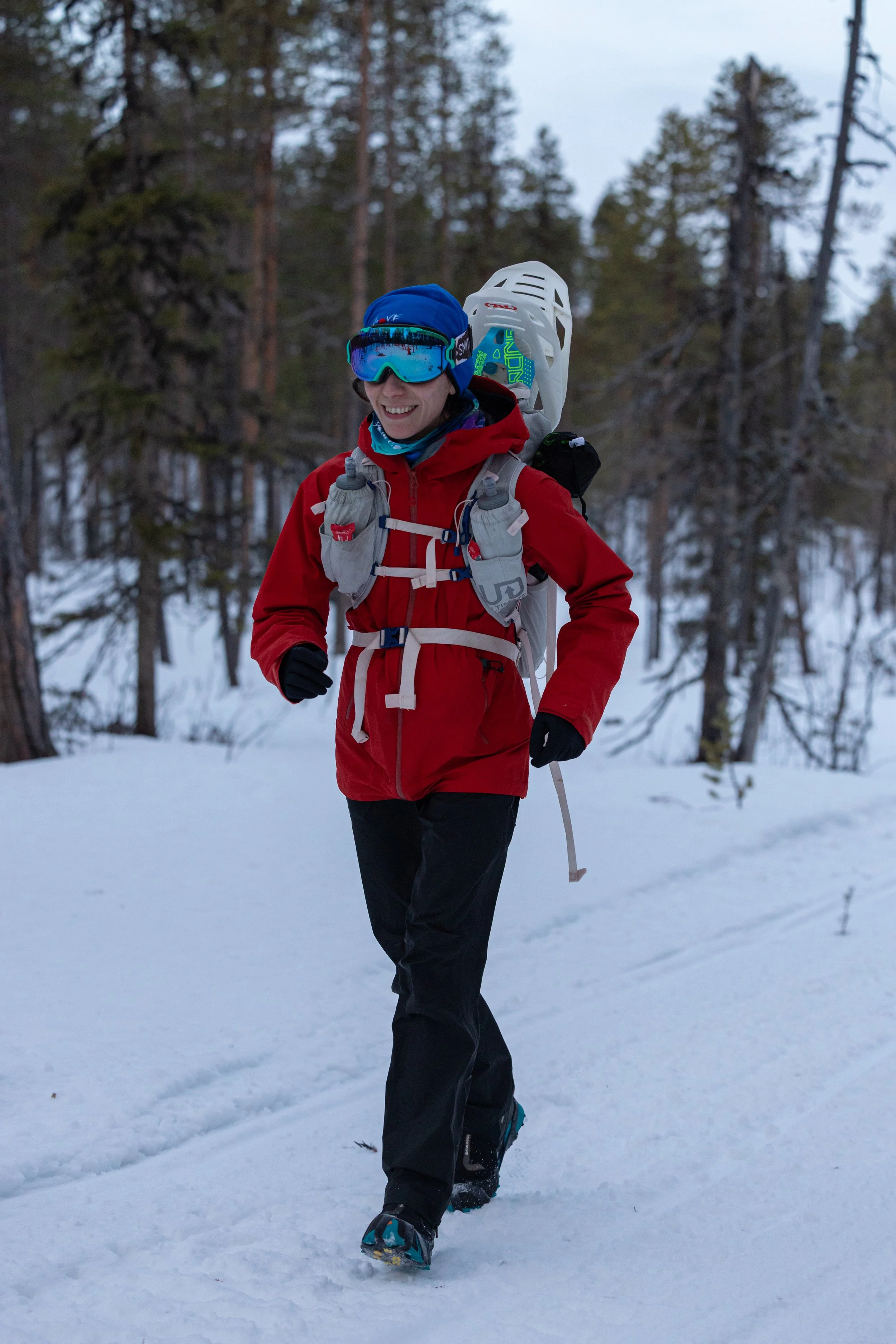 A woman hiking in the snow wearing a red jacket, black pants, blue goggles, and a backpack in a forest.