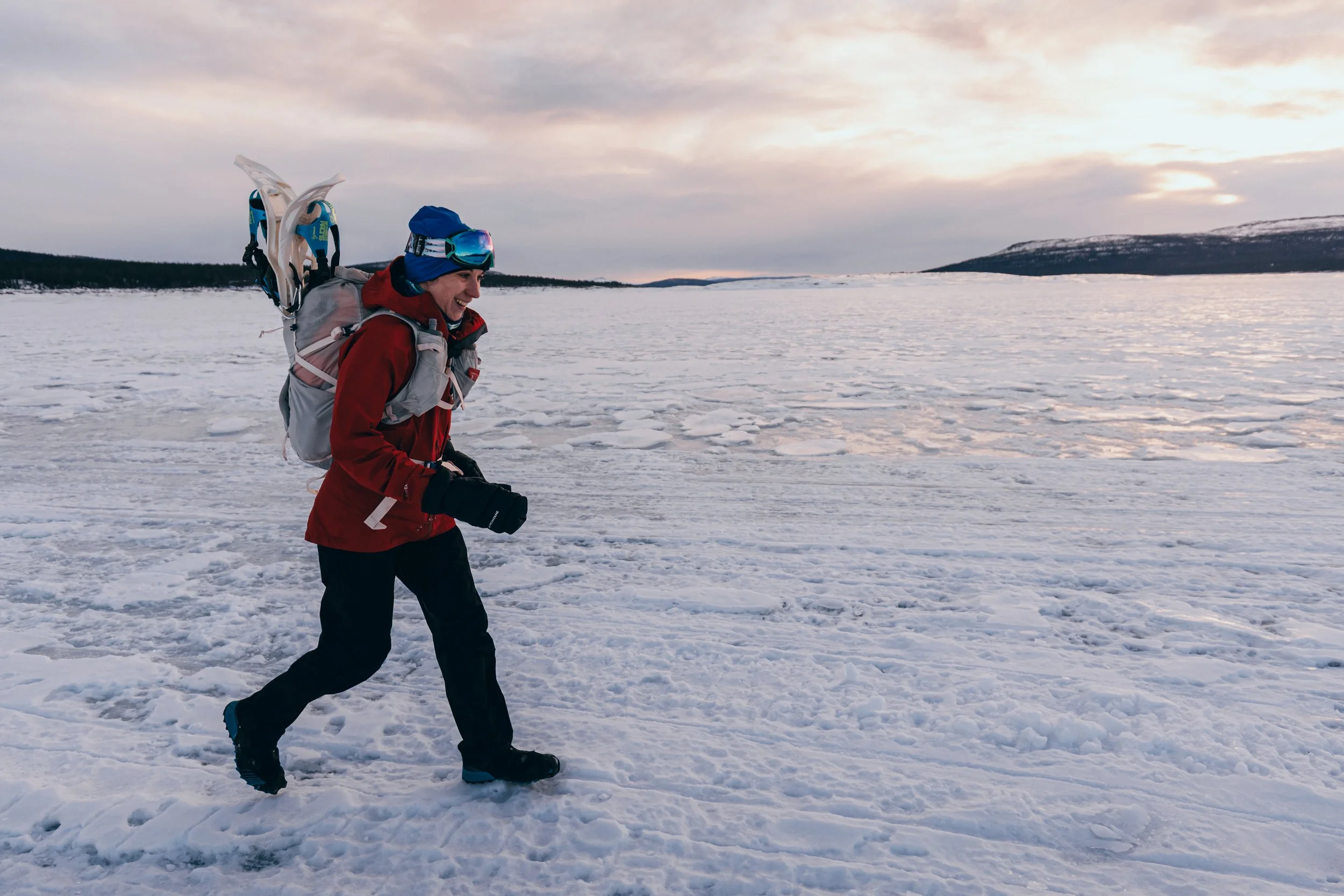 Person hiking across snow-covered landscape with snowshoes and winter gear, early morning or late afternoon sky.