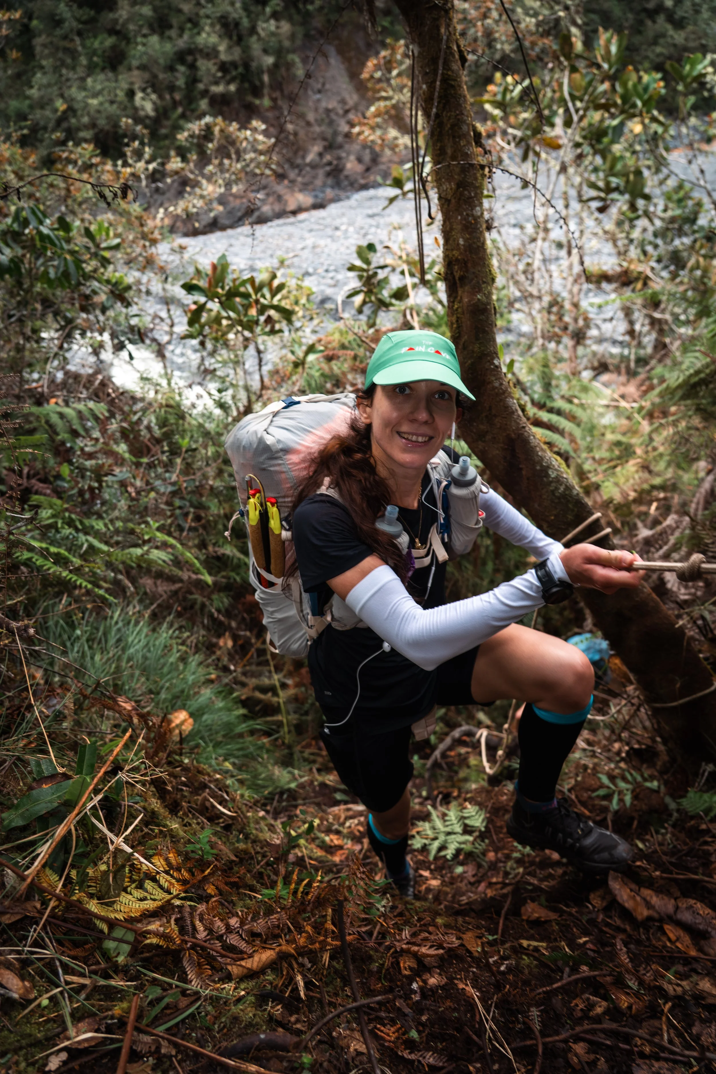 A woman wearing a teal cap, black athletic clothing, and a backpack, climbing a steep, muddy trail in a dense forest.