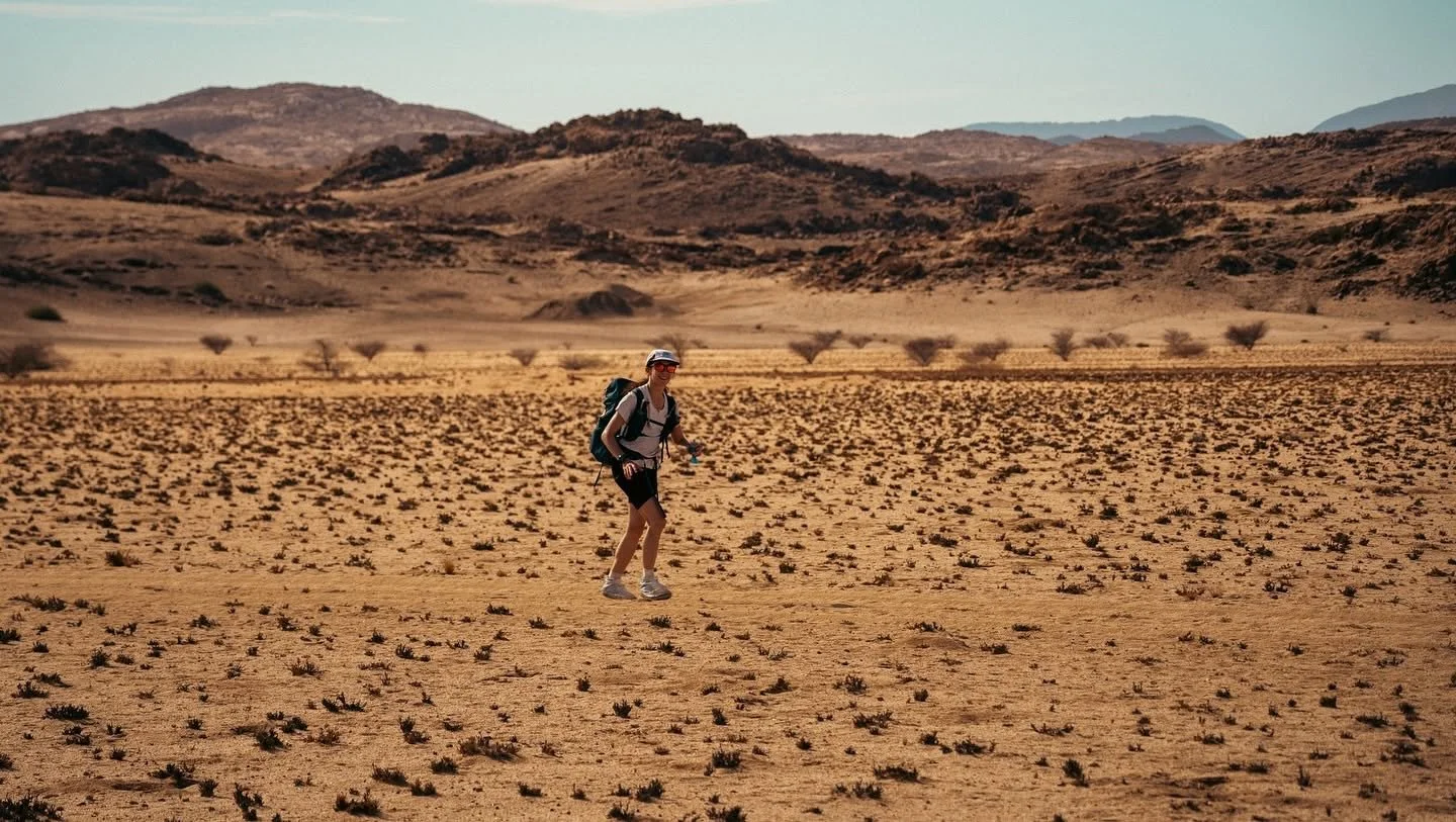 A woman hiking in a dry, desert landscape with sparse vegetation and distant mountains.