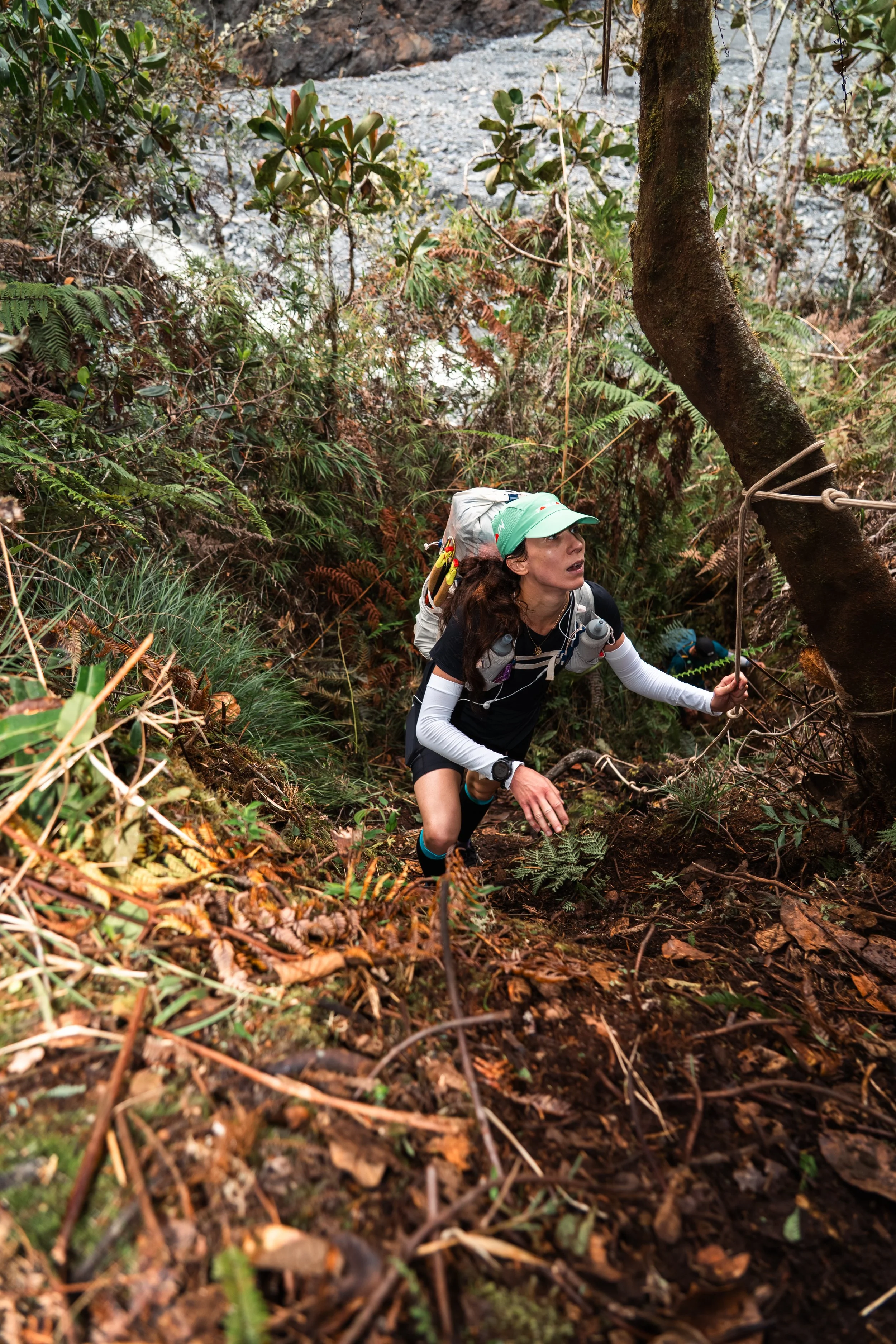 A woman in athletic gear is climbing a steep, muddy trail in a forest, holding onto a rope tied to a tree for support.