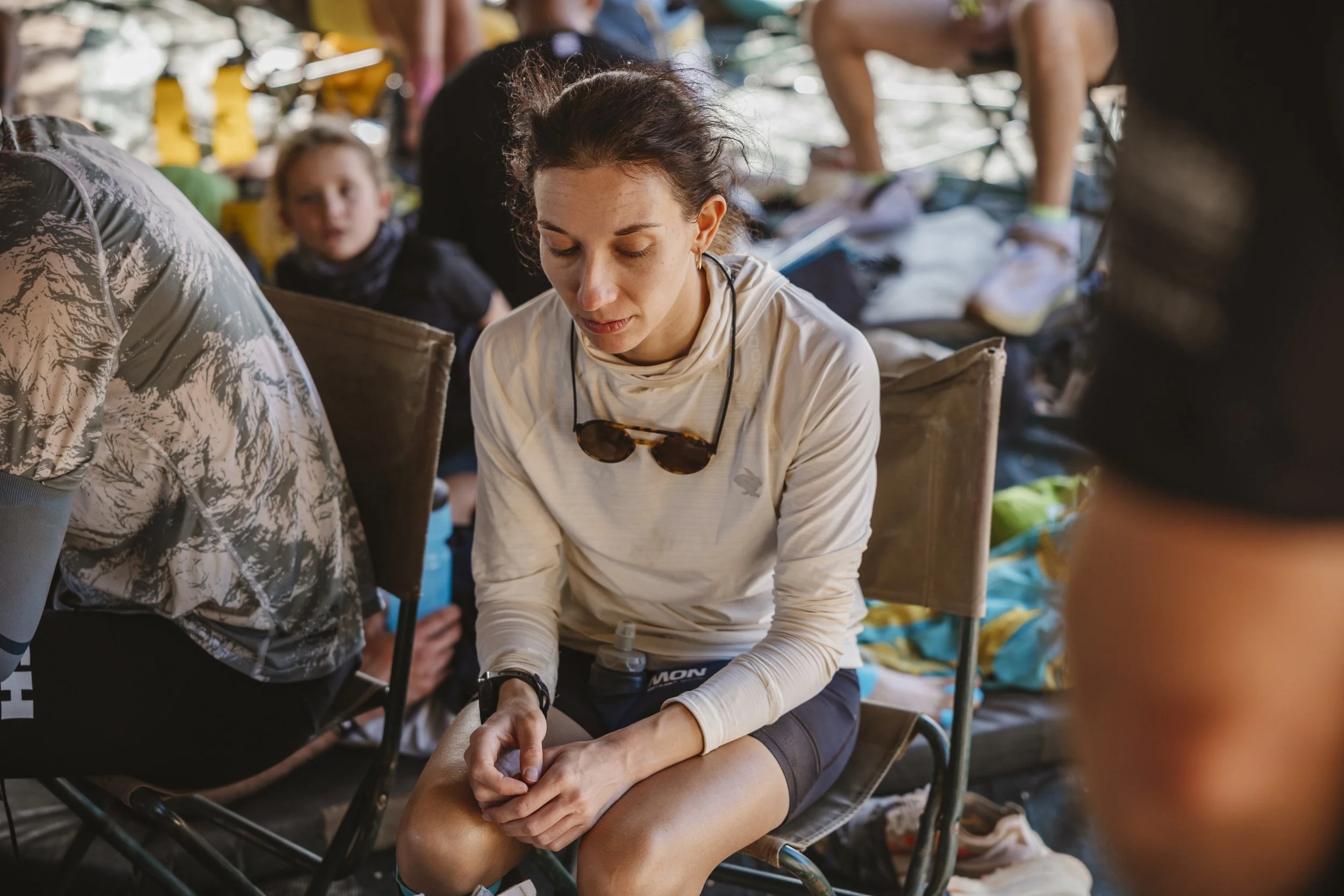 A woman with sunglasses around her neck sits in a chair with her eyes closed, surrounded by other people at an ultra marathon event, with a young girl and others in the background.