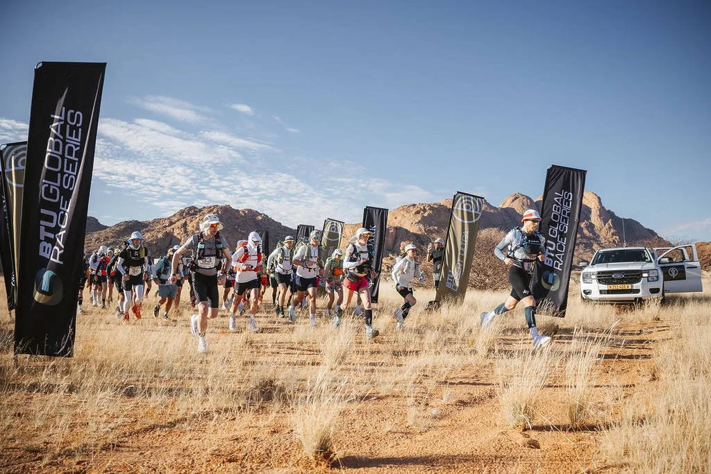 A group of runners participating in a race in a desert landscape with mountains in the background, marked by flags with the 'Big Global Race Series' logo, and a white vehicle with an open door nearby.
