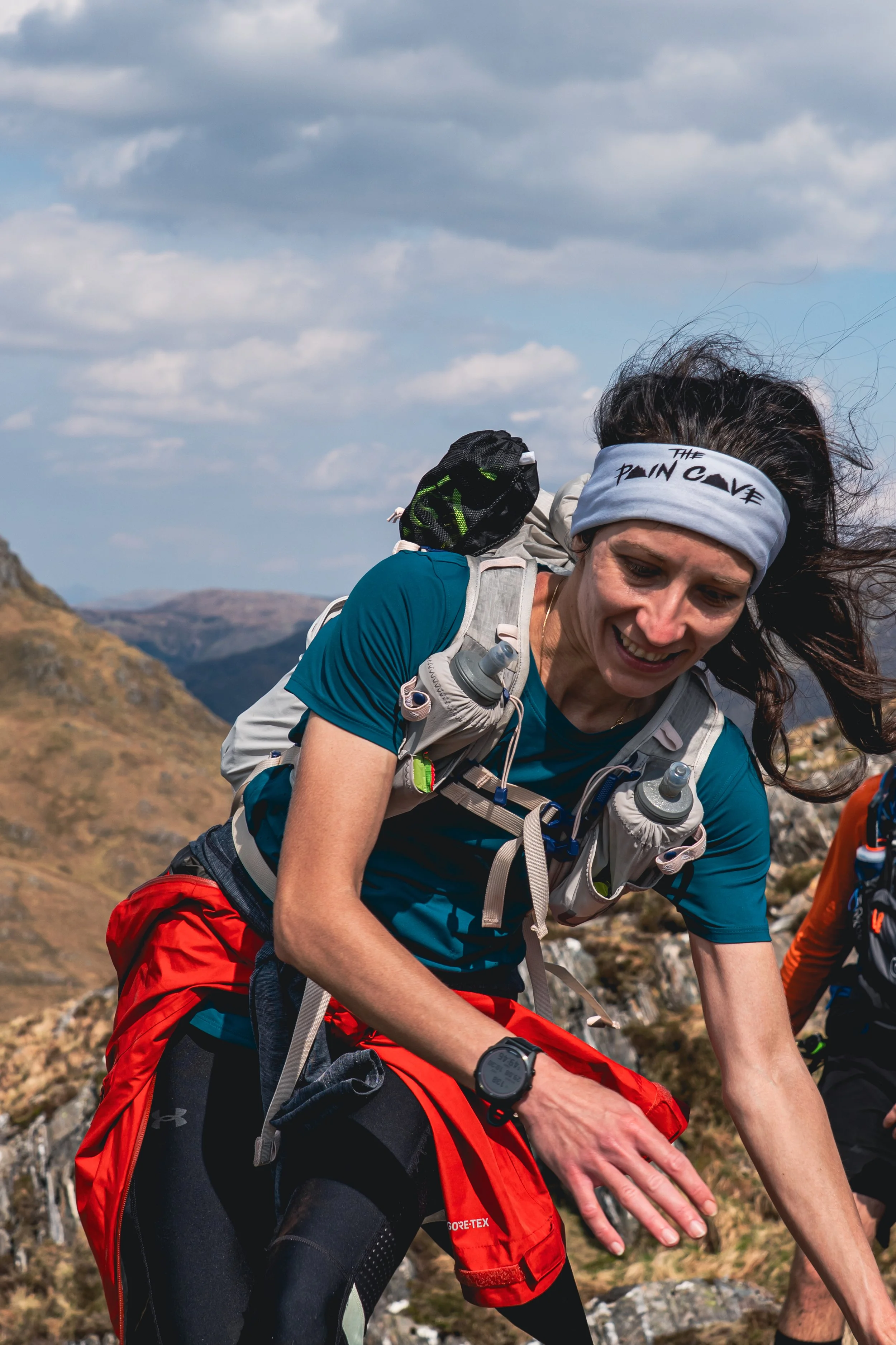 Female trail runner wearing a headband, blue shirt, and red jacket, climbing a mountain trail with backpack, in a mountainous landscape under partly cloudy sky.