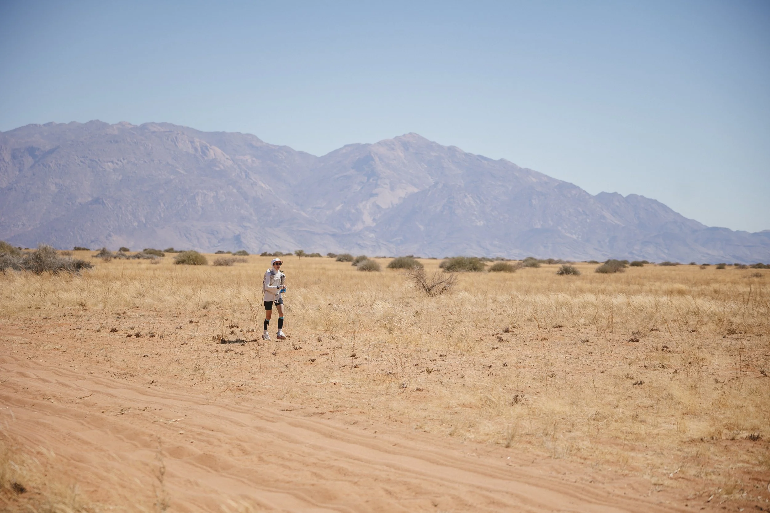 A person in athletic clothing and a hat walking in a dry desert landscape with mountains in the background.