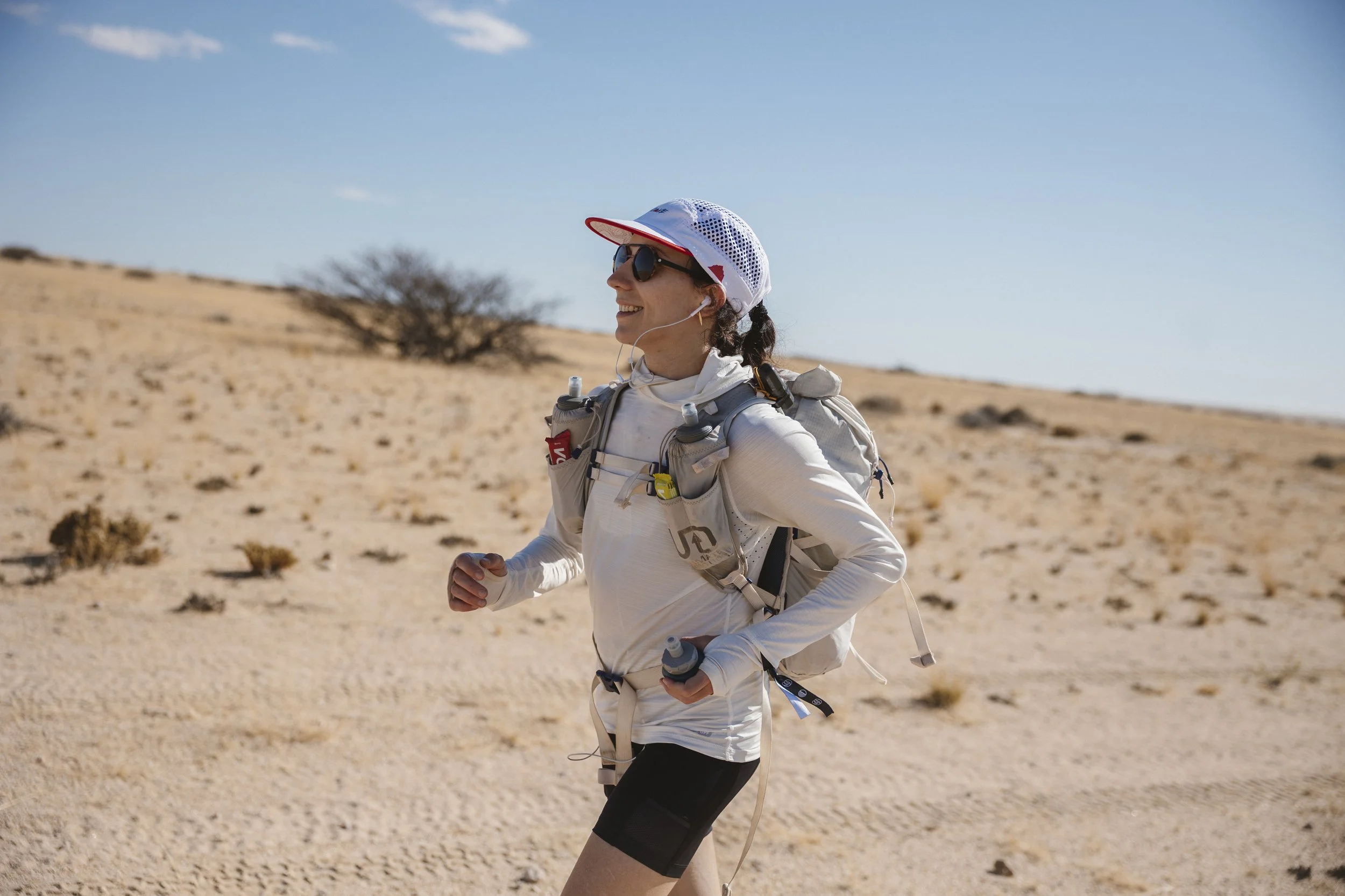 A woman running in a desert landscape, dressed in athletic attire with a white long-sleeve shirt, black shorts, sunglasses, a cap, and a hydration backpack.
