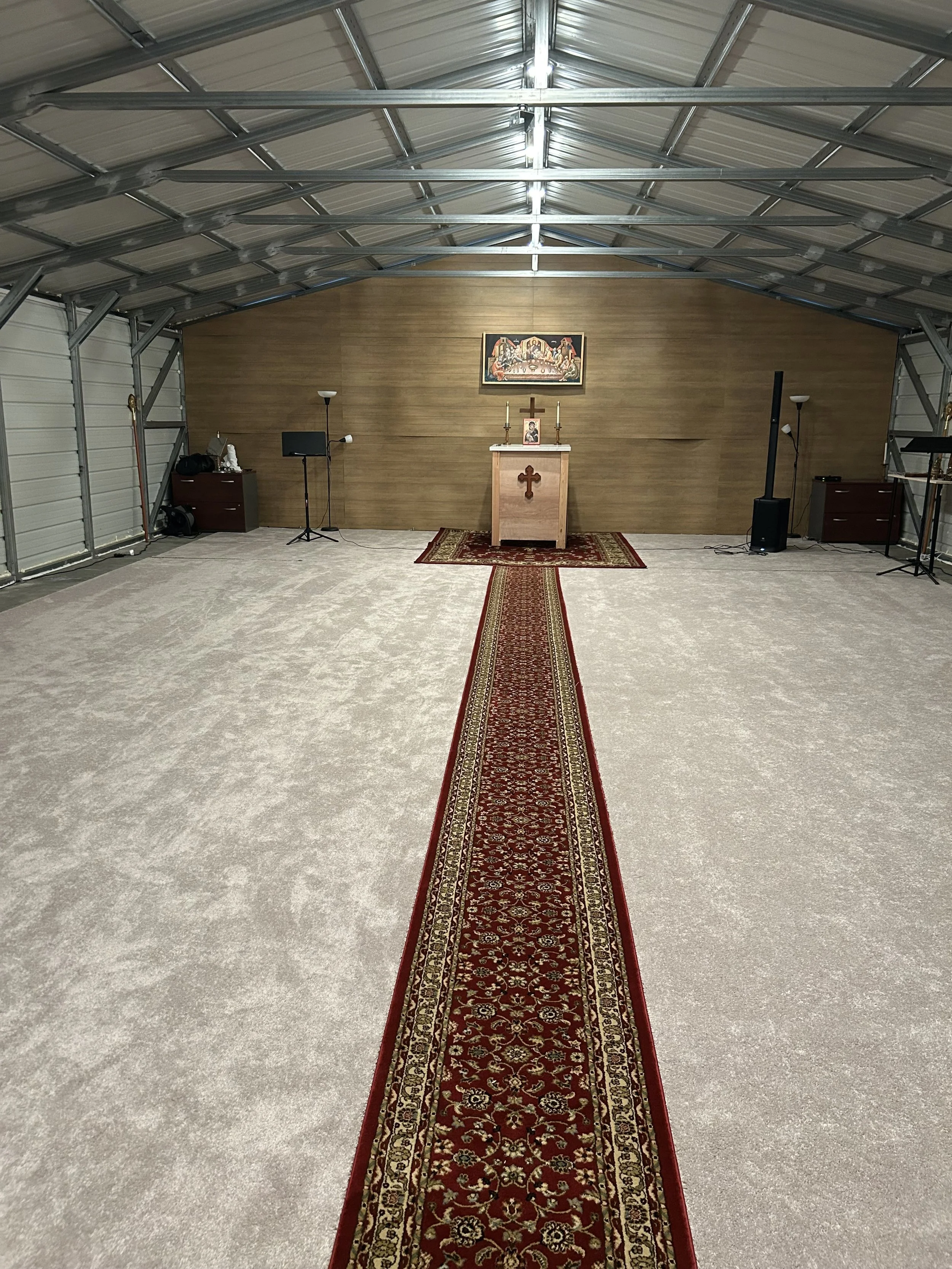 Empty church altar area with a red patterned carpet runner, wooden altar table with a cross, religious art above, and church-related items on the sides, inside a metal structure with a curved ceiling.