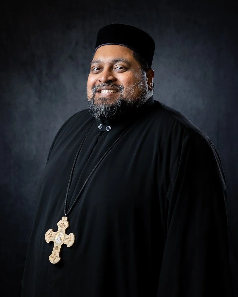A man in religious attire, wearing a black robe and a large gold cross necklace, smiling against a dark background.