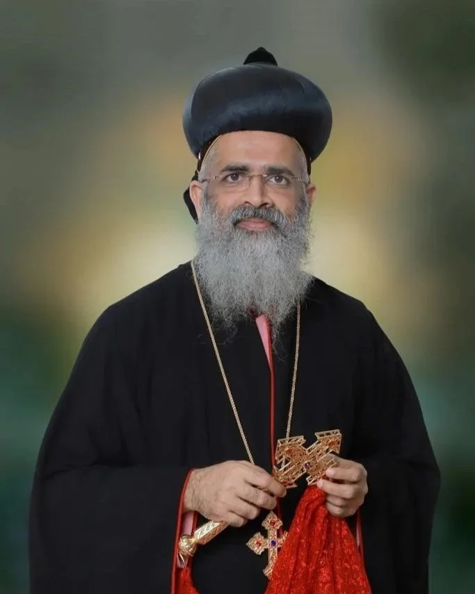 A man dressed in traditional religious attire, wearing a black robe and a large black headpiece, holding religious symbols, with a long gray beard, standing against a blurred green background.