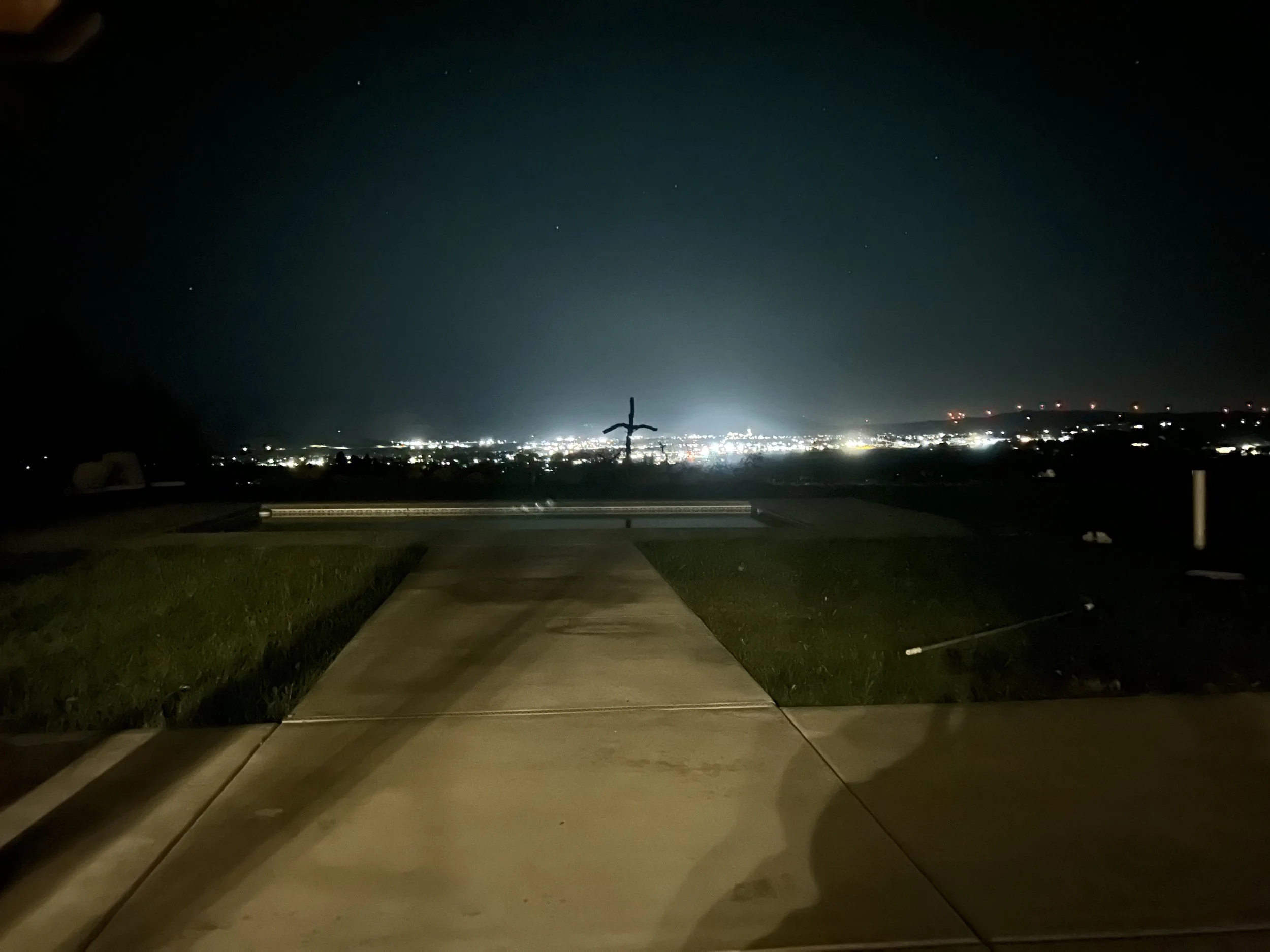 Nighttime view from a backyard with a sidewalk, grassy area, and a city skyline in the distance illuminated with lights, with a prominent telephone pole in the middle.