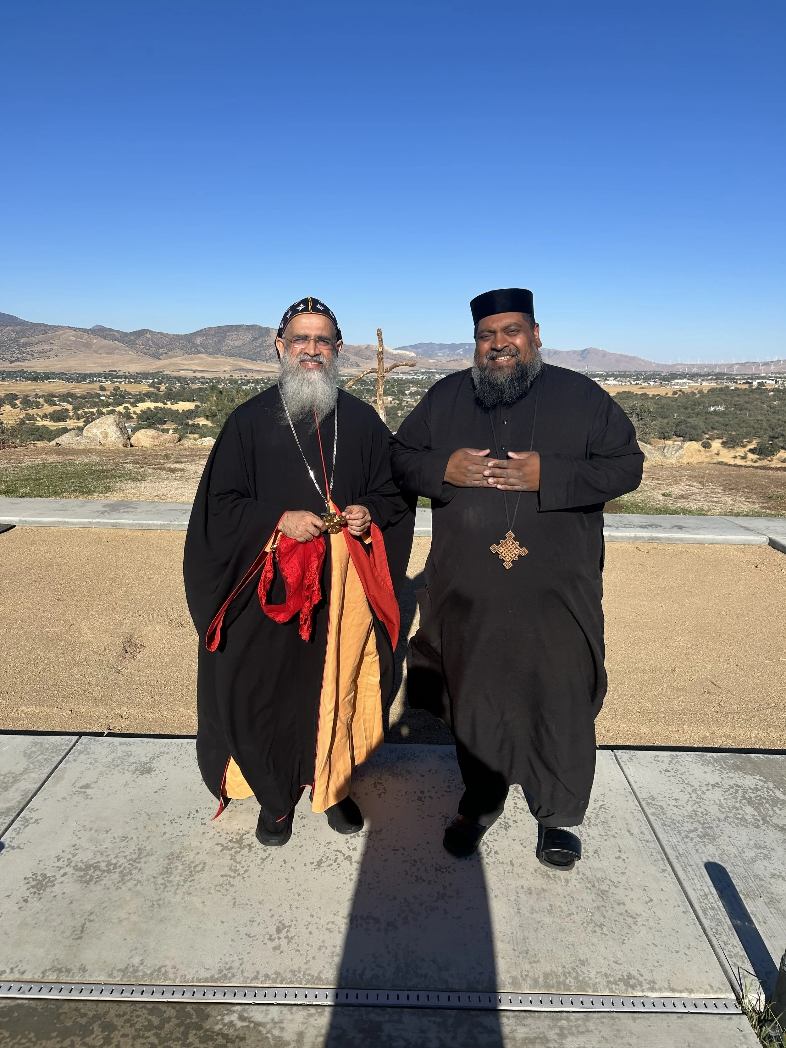 Two men dressed in religious robes standing outdoors with a scenic landscape of mountains and a clear blue sky in the background.