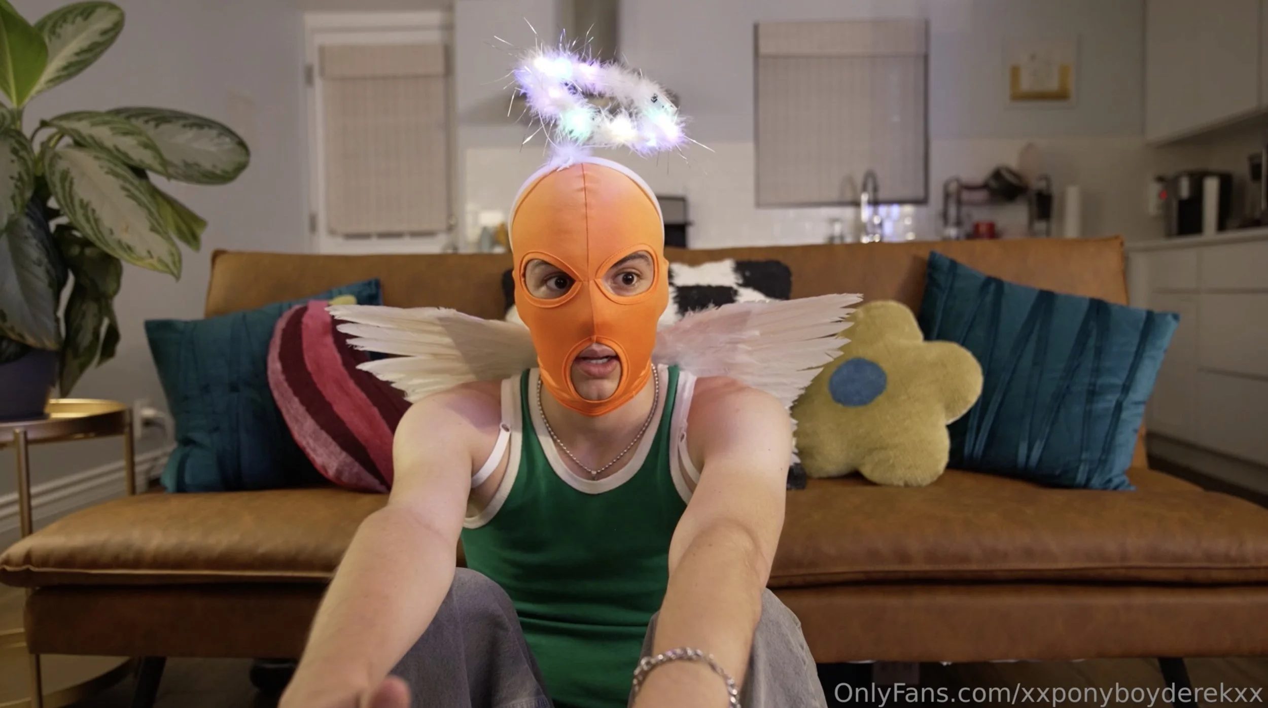 A person wearing an orange ski mask with cutouts for the eyes, nose, and mouth, along with white fairy wings, seated on a brown couch in a living room. They have a green tank top, a beaded bracelet, and a small chain necklace. The background includes cushions, a potted plant, and kitchen area.