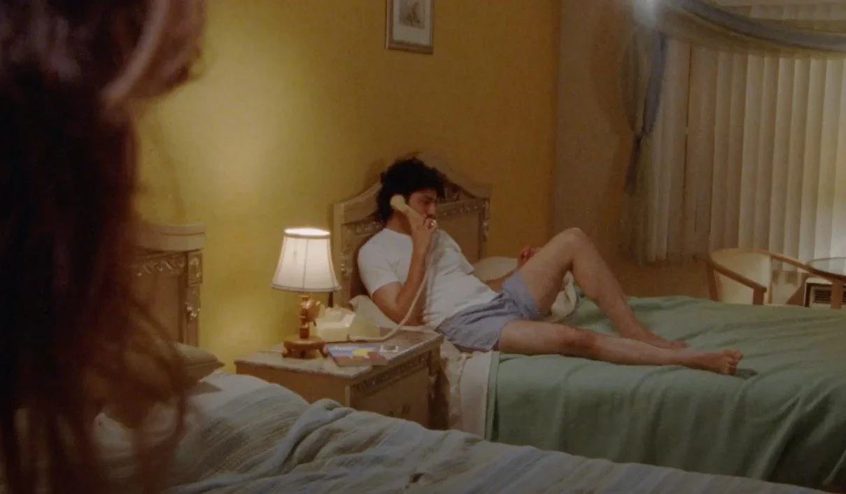 A man lying on a hotel bed talking on a landline phone, wearing a white t-shirt and shorts, with a woman partially visible in the foreground.