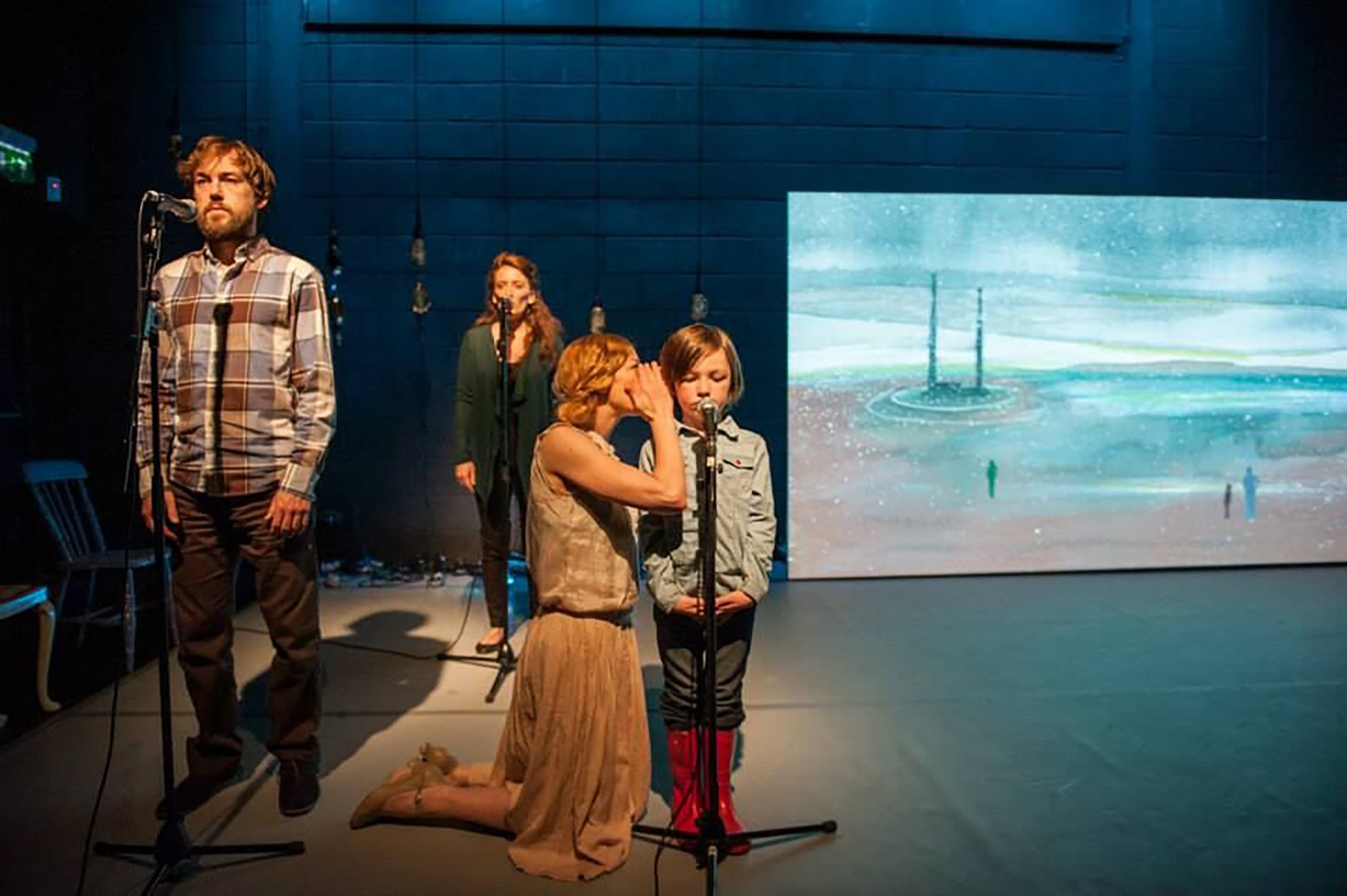 A theatrical performance with four actors on stage. One woman is kneeling and whispering to a young boy at a microphone. A man stands to the left speaking into a microphone, and a woman in the background is standing near a microphone. Behind them, a large screen displays an animated scene of a fountain in a rainy landscape.